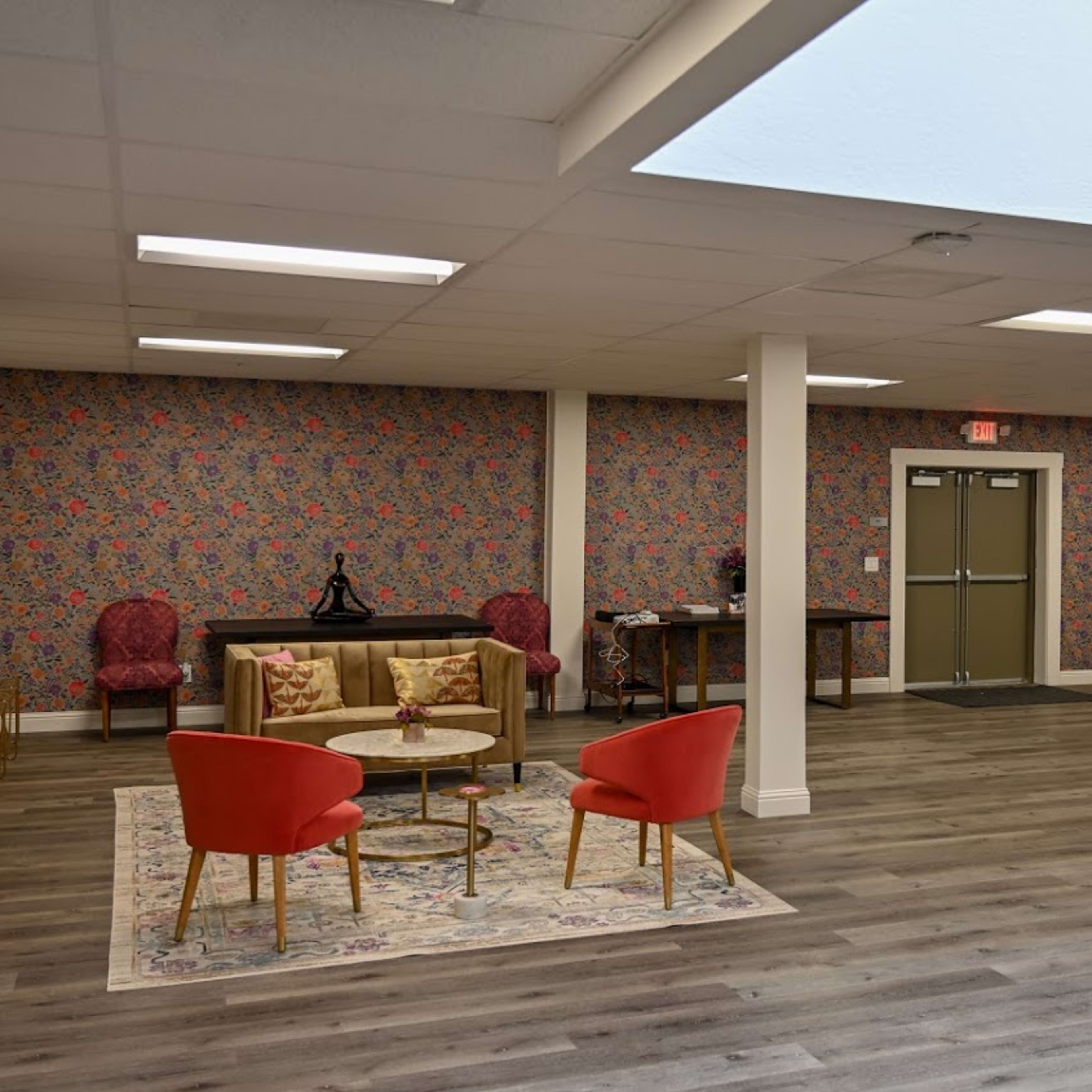 The interior of a spacious room features a mixture of seating arrangements, including red chairs and a long dining table, with decorative wallpaper and natural lighting from a skylight.