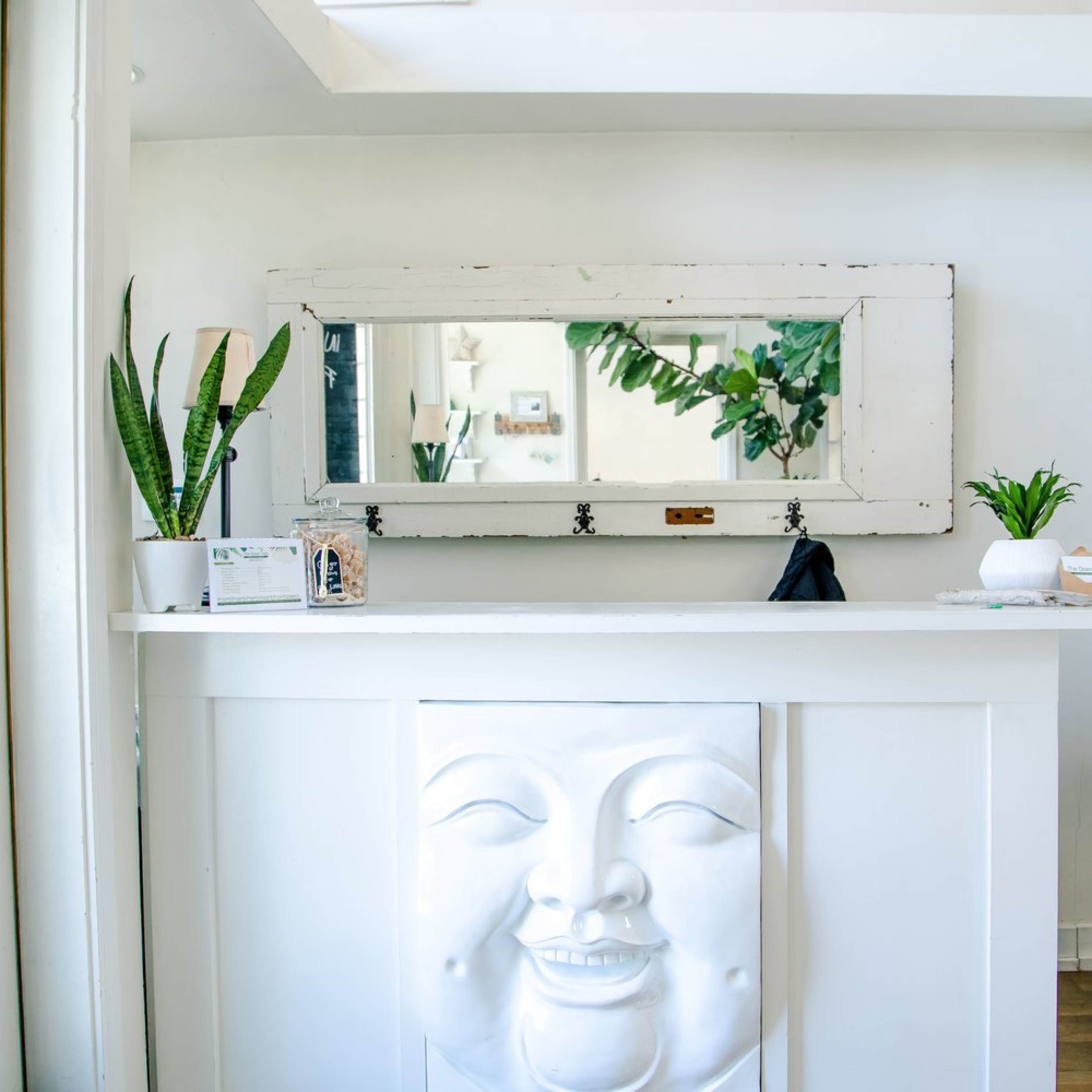 A reception area featuring a white counter with a smiling face relief and a large mirror above it, flanked by potted plants.