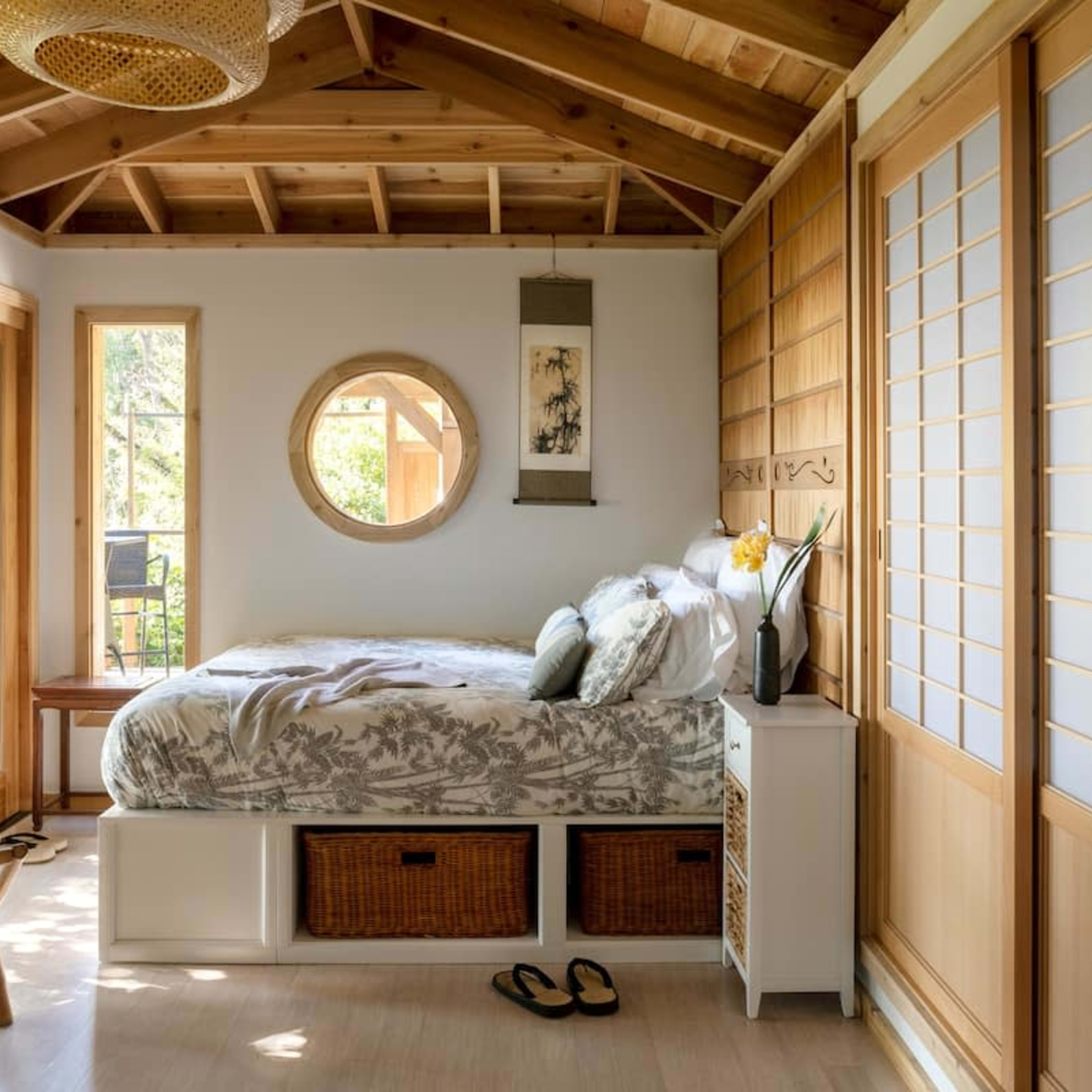 A cozy bedroom with a bed, wooden accents, large windows, and a small dining area featuring a chair and a table.