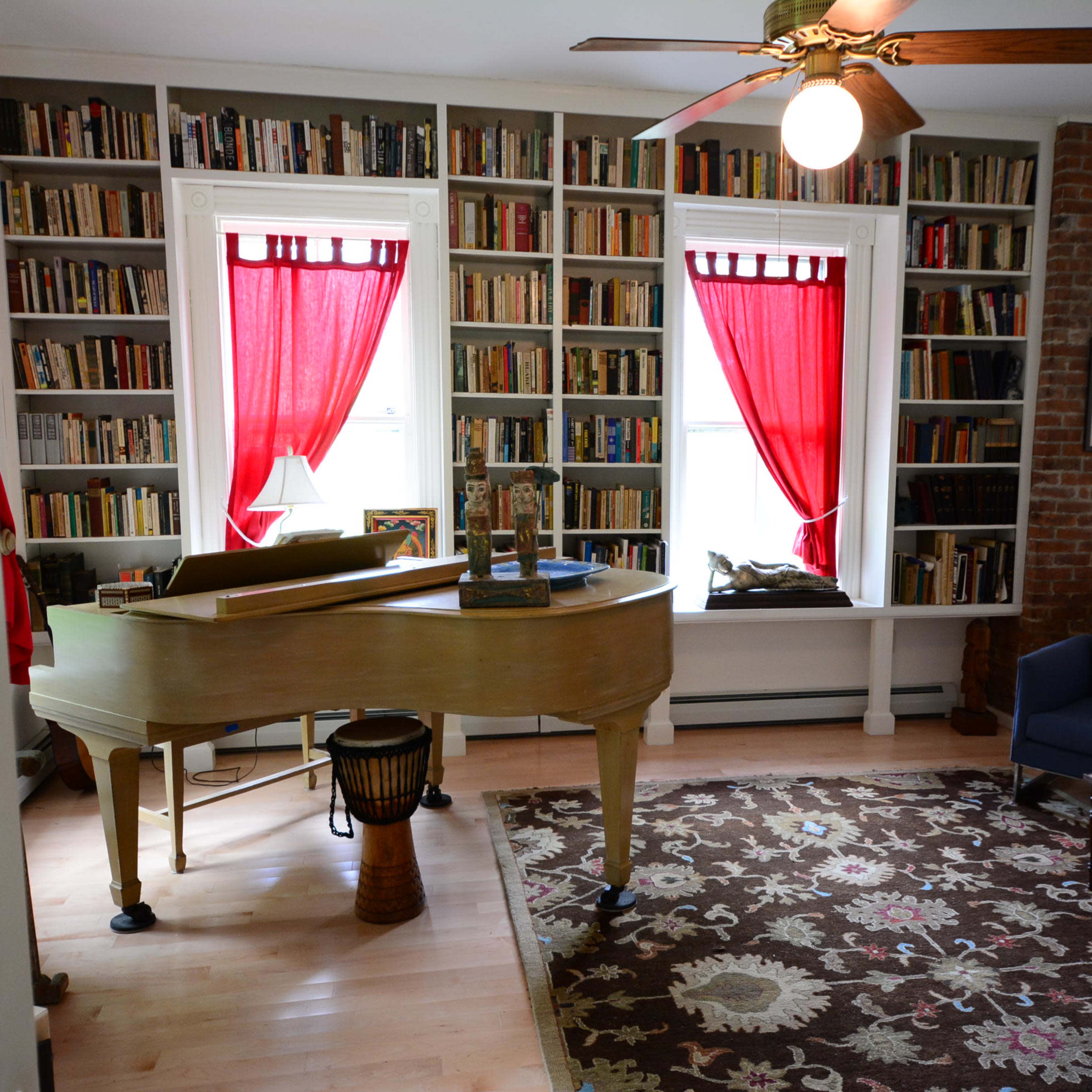 A room with a grand piano, a blue armchair, and bookshelves filled with books, accented by red curtains at the windows.