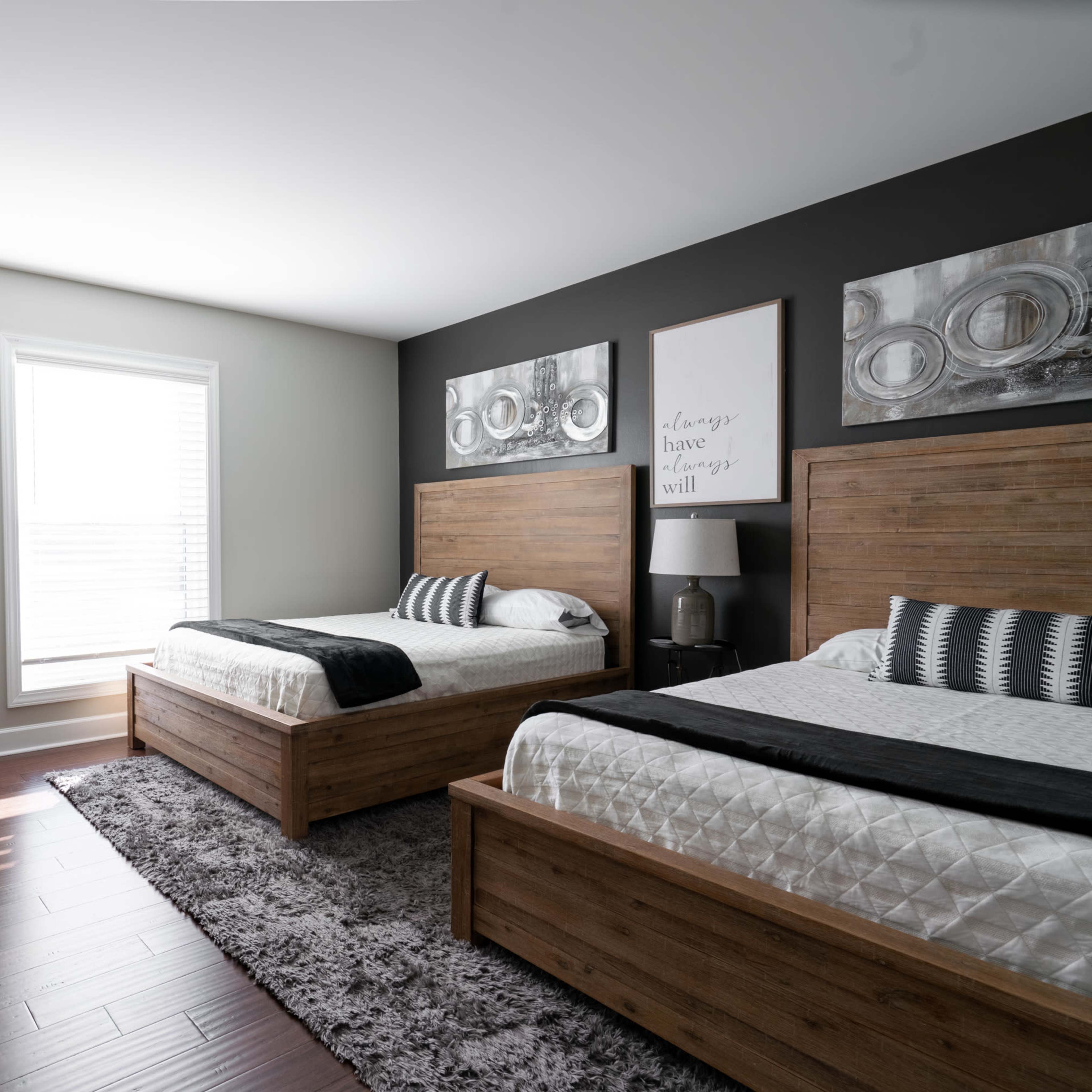 A well-lit bedroom featuring two wooden beds, a gray area rug, a mirror, a small armchair, and wall art above the beds.