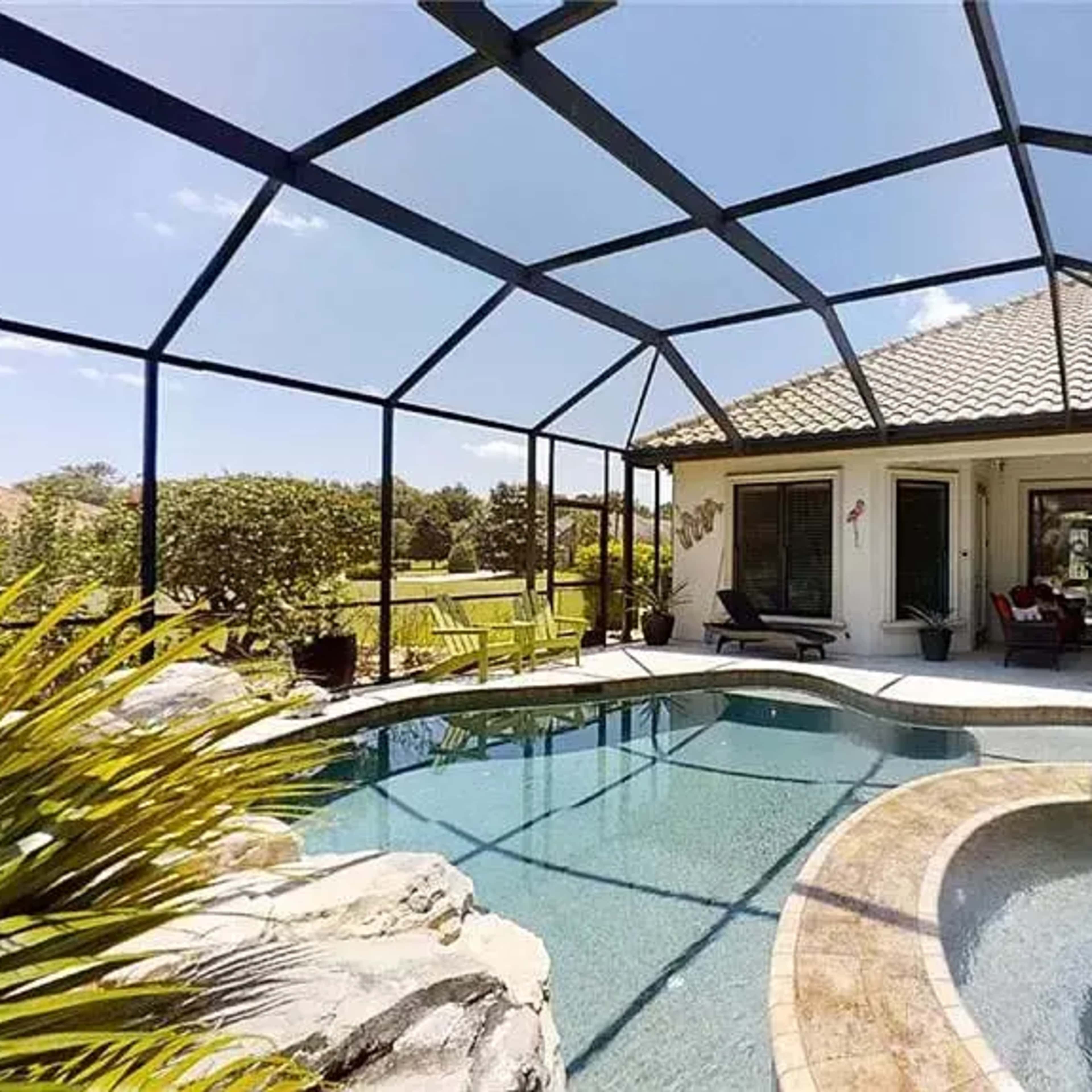 A screened-in patio area with a curved swimming pool and a spa, set against a backdrop of green landscaping and a house with a tiled roof.