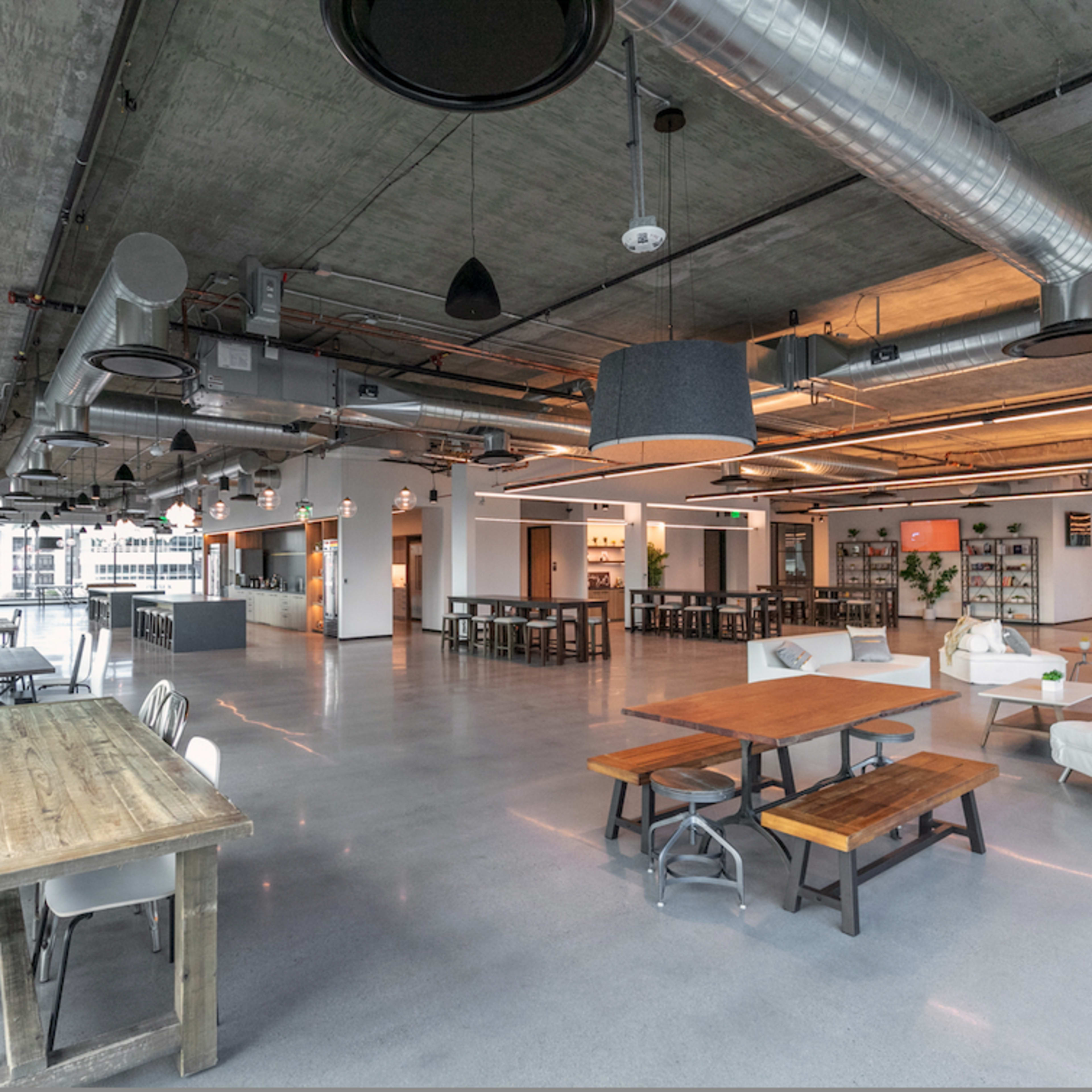 A spacious, modern industrial-style office interior with large windows, concrete walls, and various seating arrangements, including tables and couches.