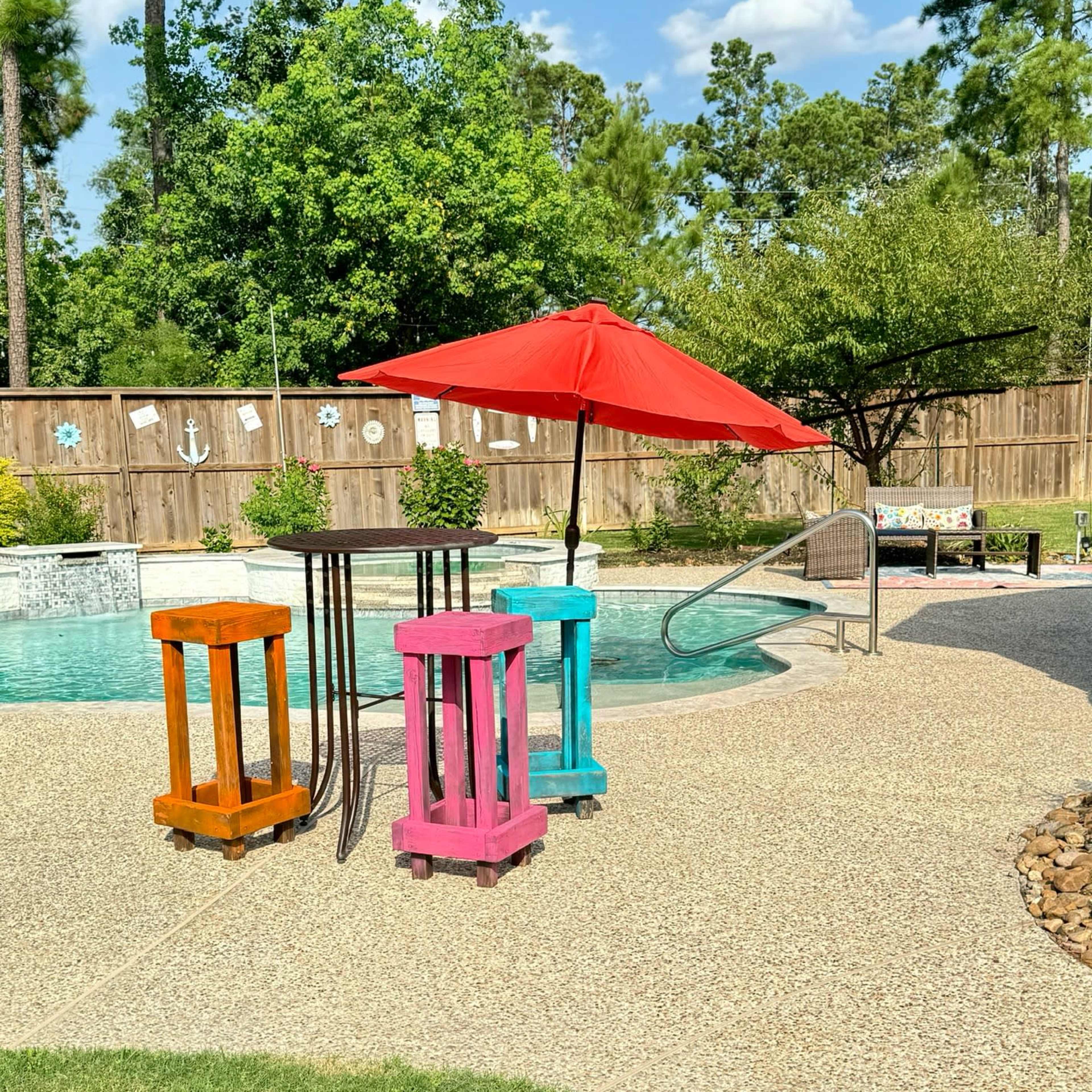 The scene features colorful tables in front of a pool, with a red umbrella providing shade, surrounded by grass and trees.