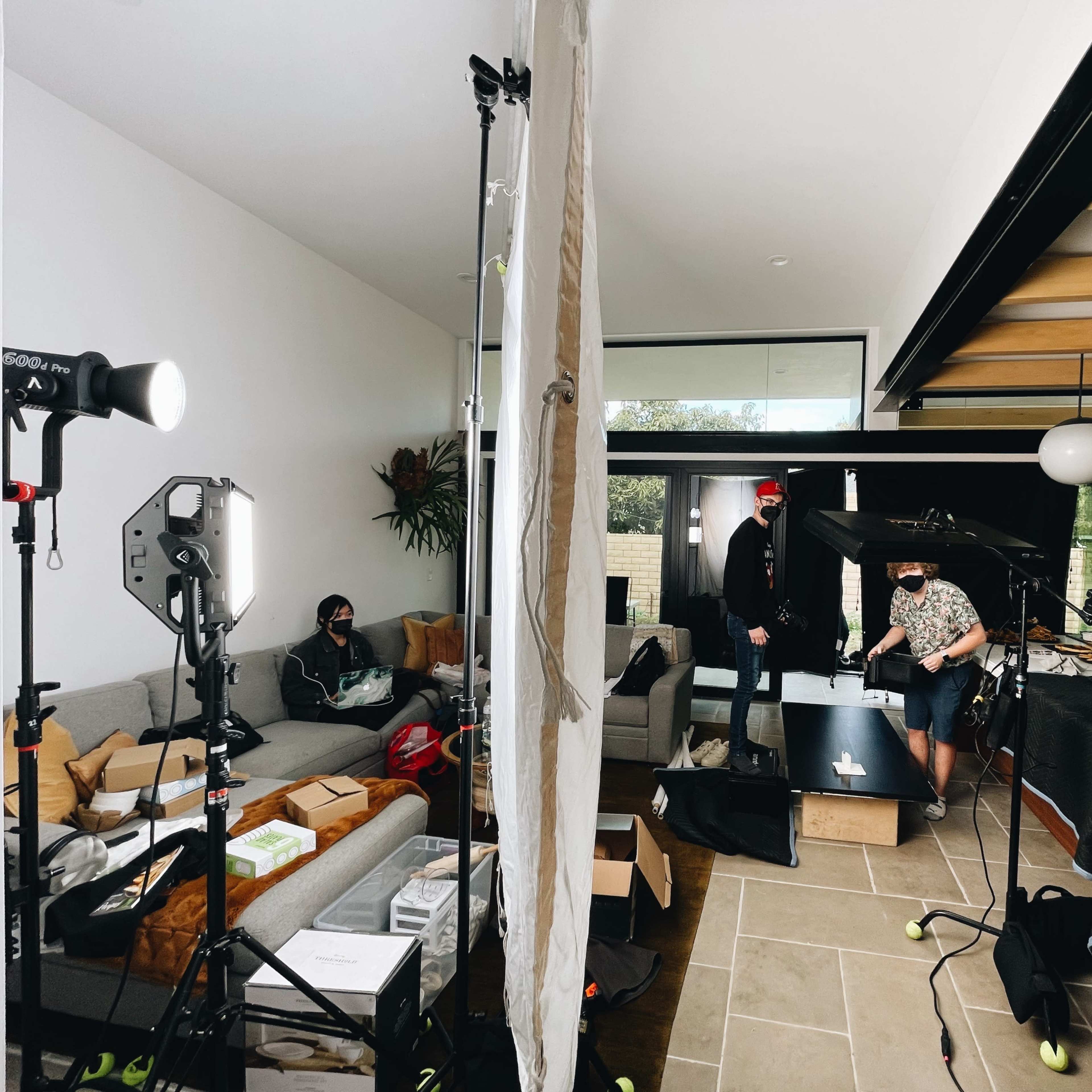 A cluttered living room set up for a photoshoot, featuring various equipment, two people wearing masks, and bright lighting.