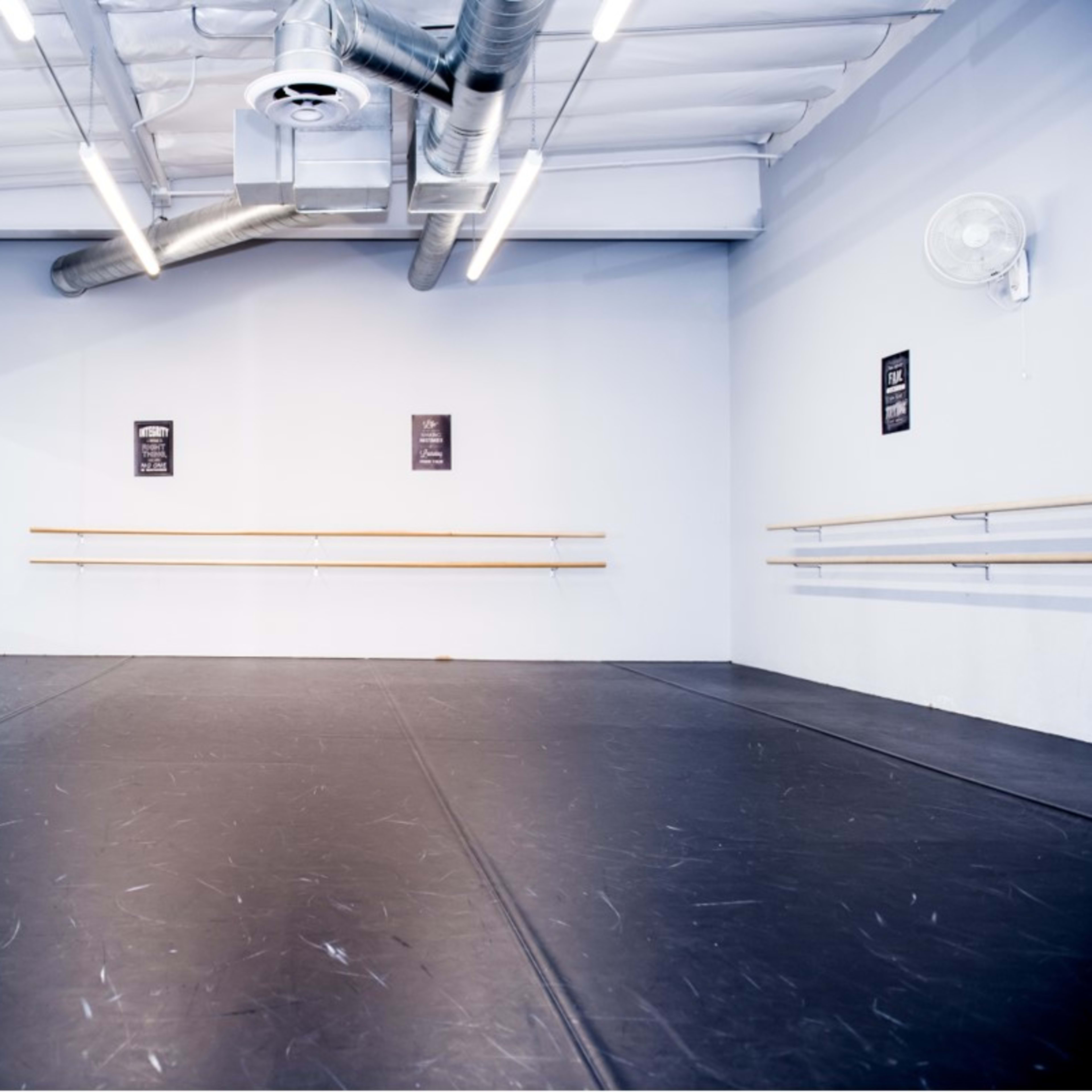 A spacious dance studio with a wooden ballet bar, mirrors on one wall, and a black rubber floor.