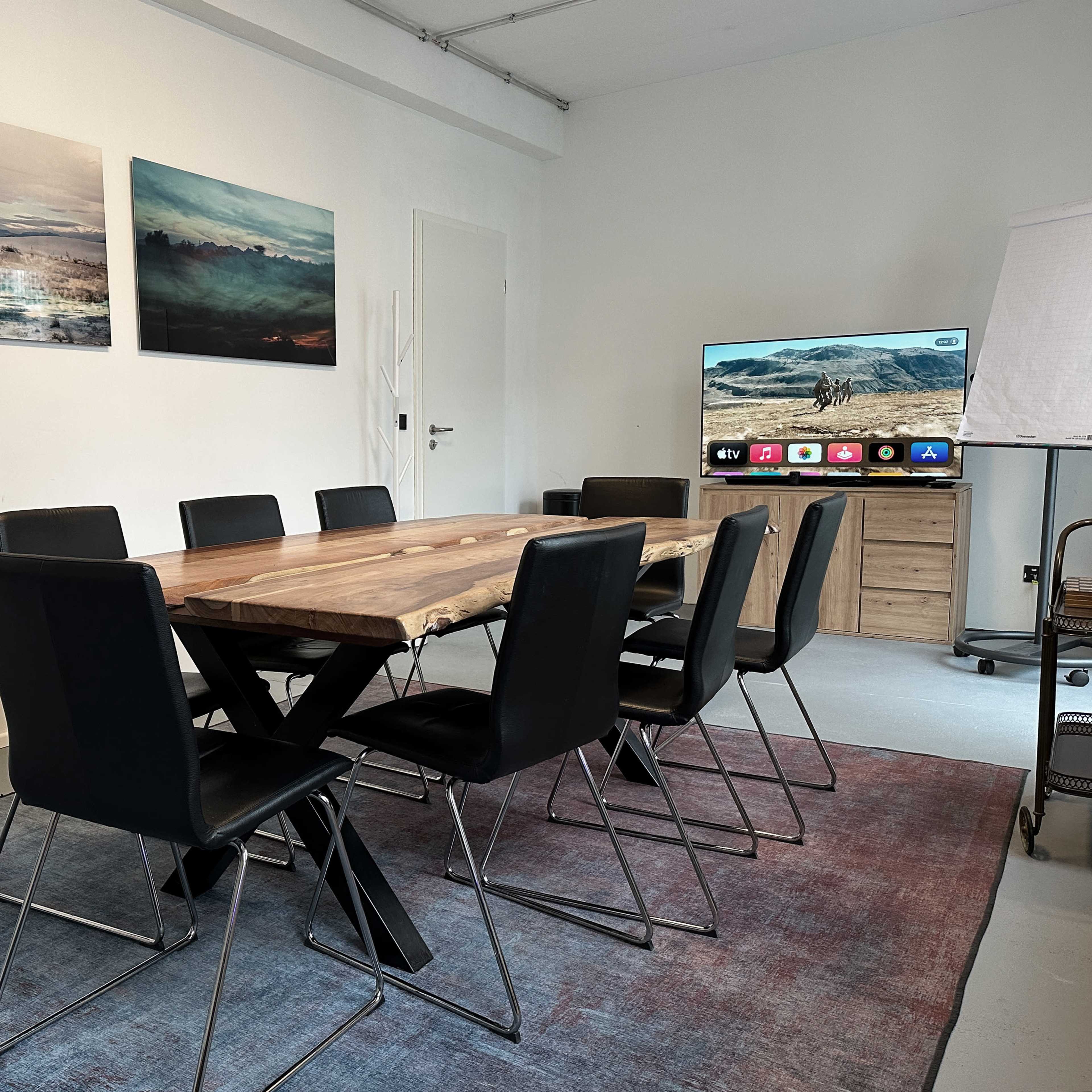 A modern conference room features a wooden table surrounded by black chairs, with two landscape paintings on the wall and a television on a stand.