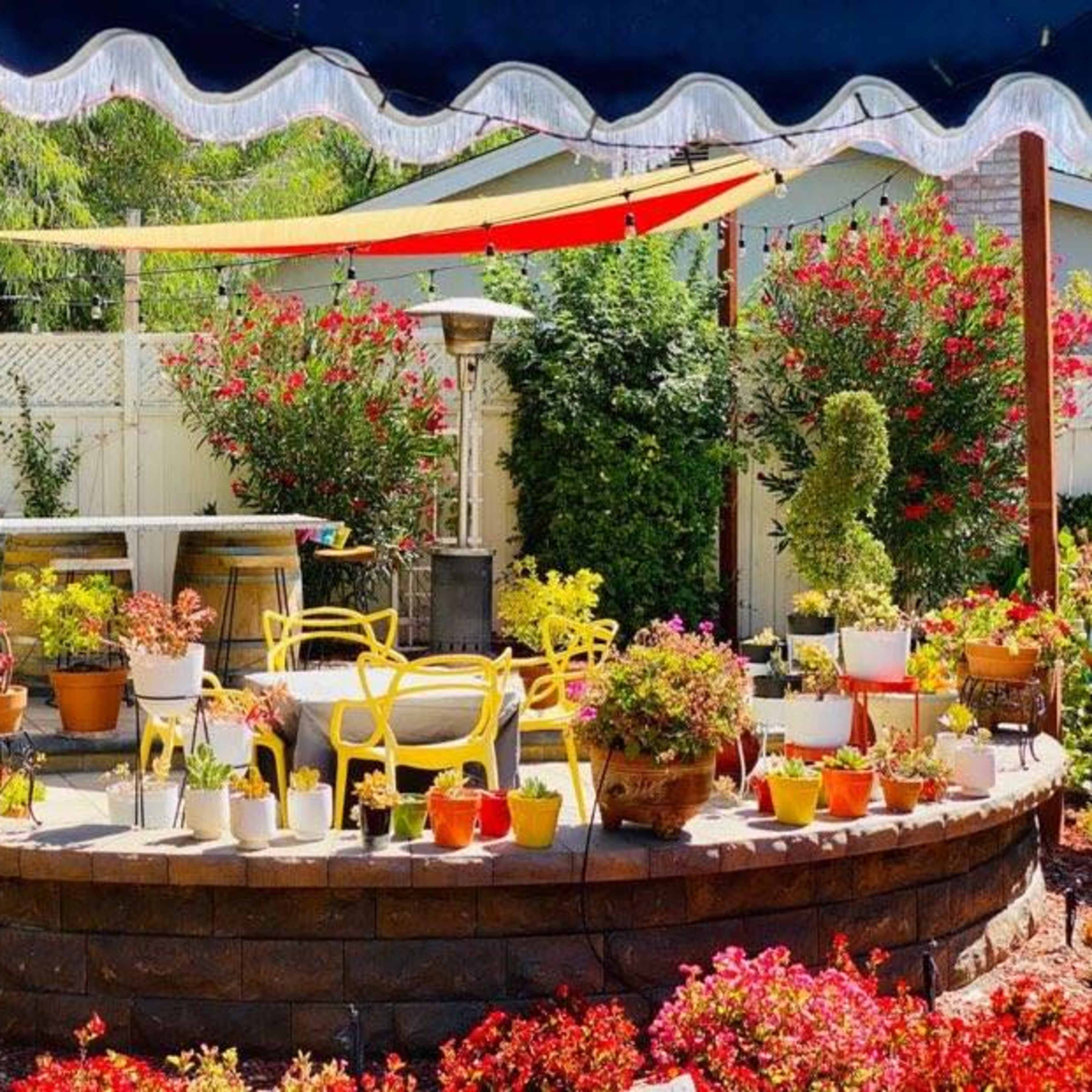 A colorful garden patio features yellow chairs and various potted plants, surrounded by flowering shrubs and a shaded area.