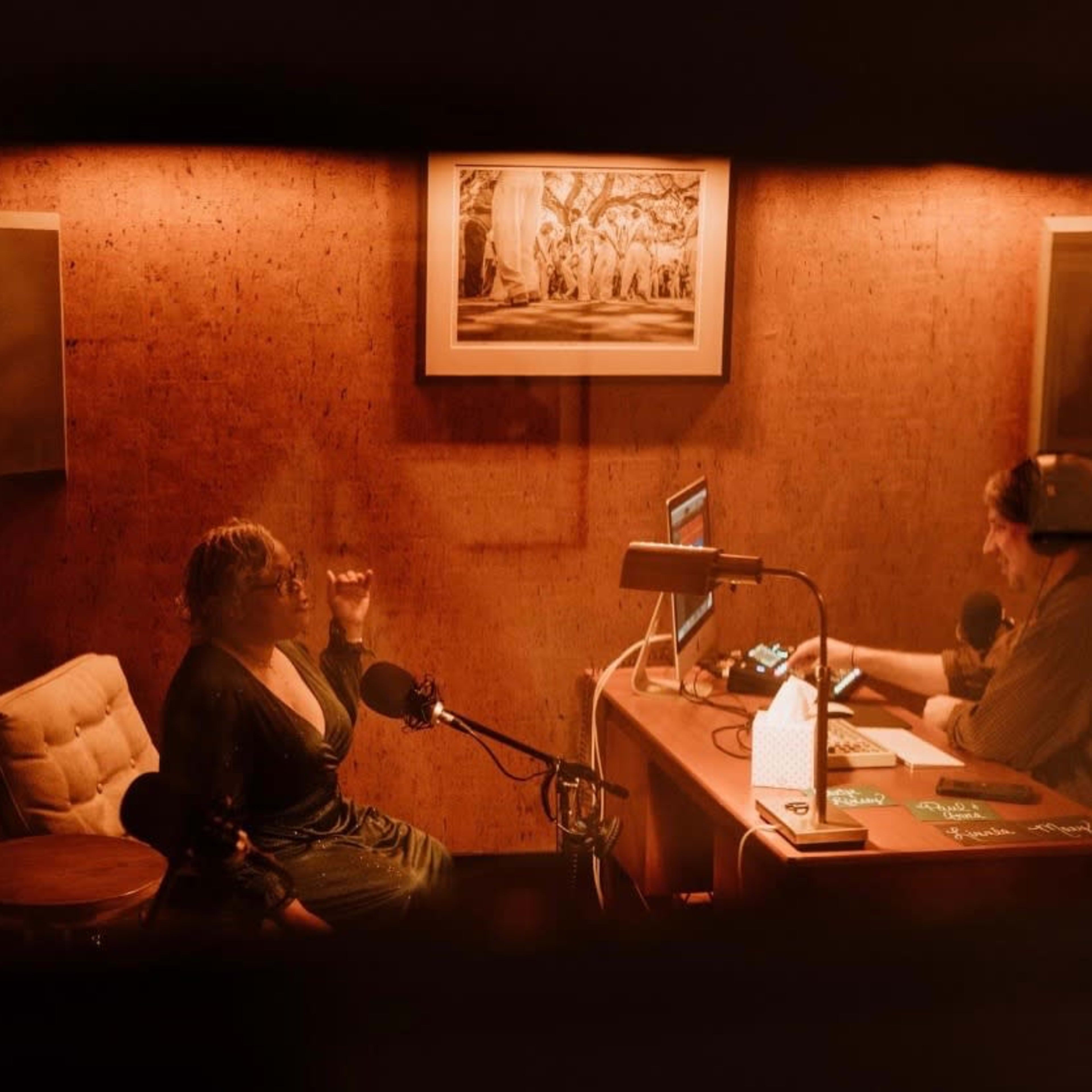 A woman speaks animatedly while seated in a recording studio, as a man listens and operates equipment at a nearby desk.