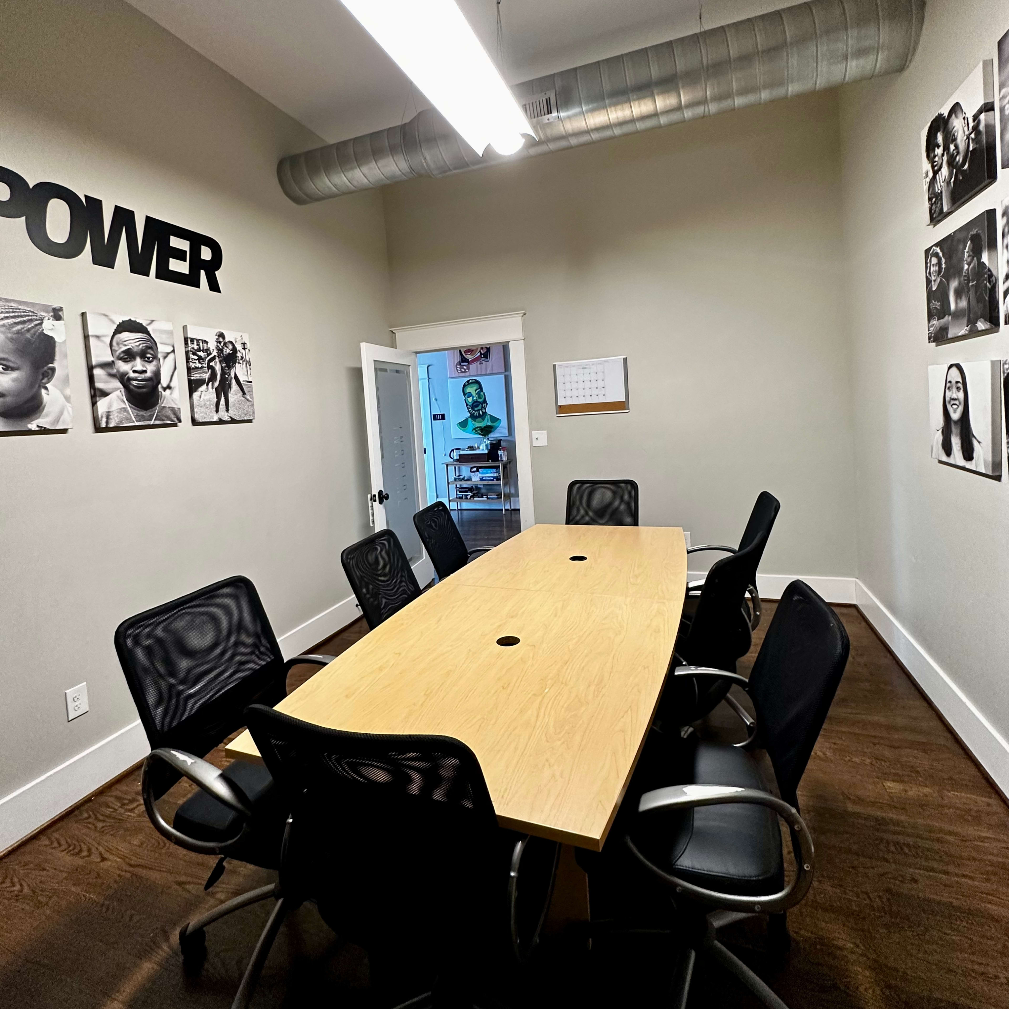 A conference room features a long wooden table surrounded by black mesh chairs, with black-and-white photographs on the walls and a door leading to another room in the background.