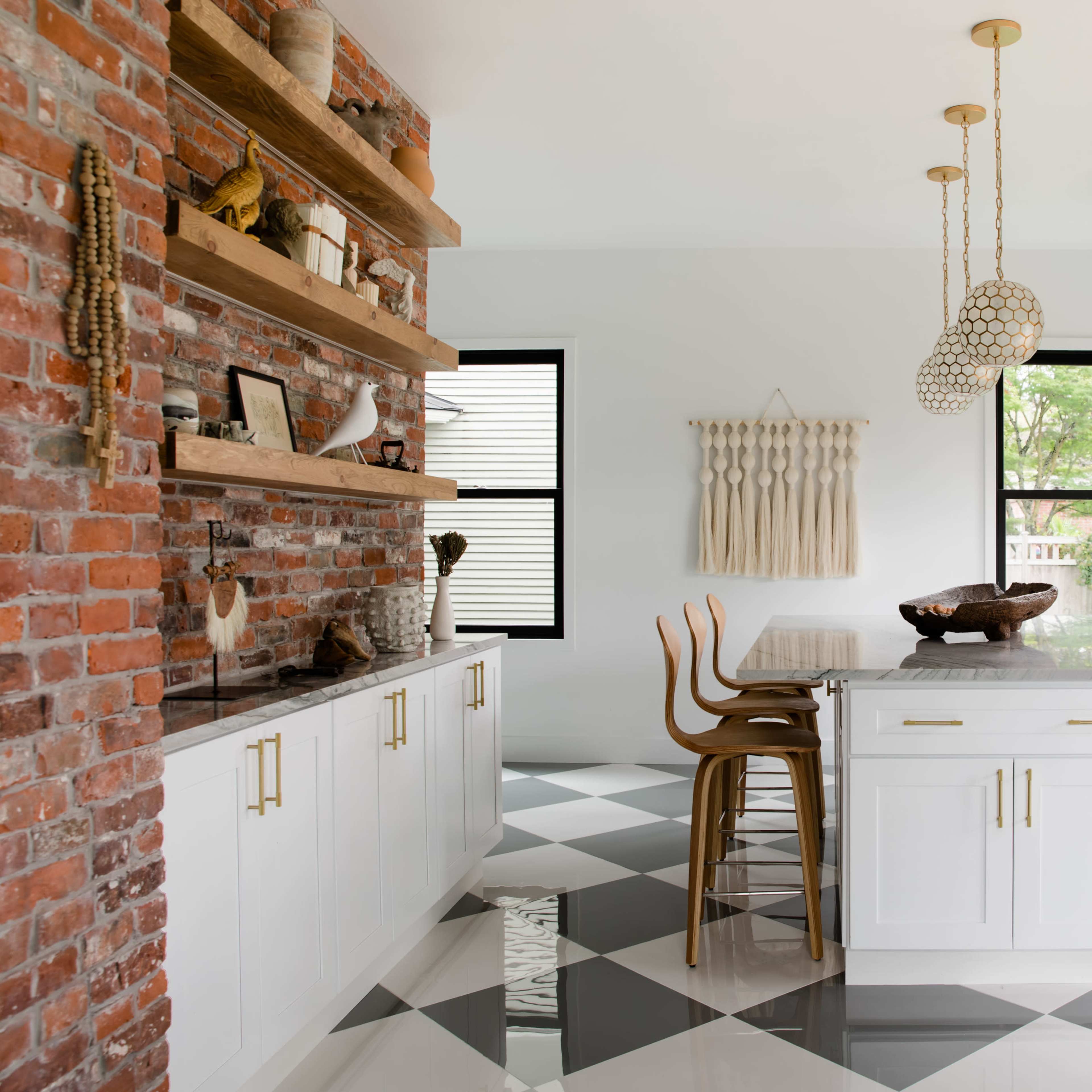 A modern kitchen features a brick wall, open shelving with decorative items, a central island with a wooden bowl, and a dining table surrounded by wooden chairs on a checkered floor.