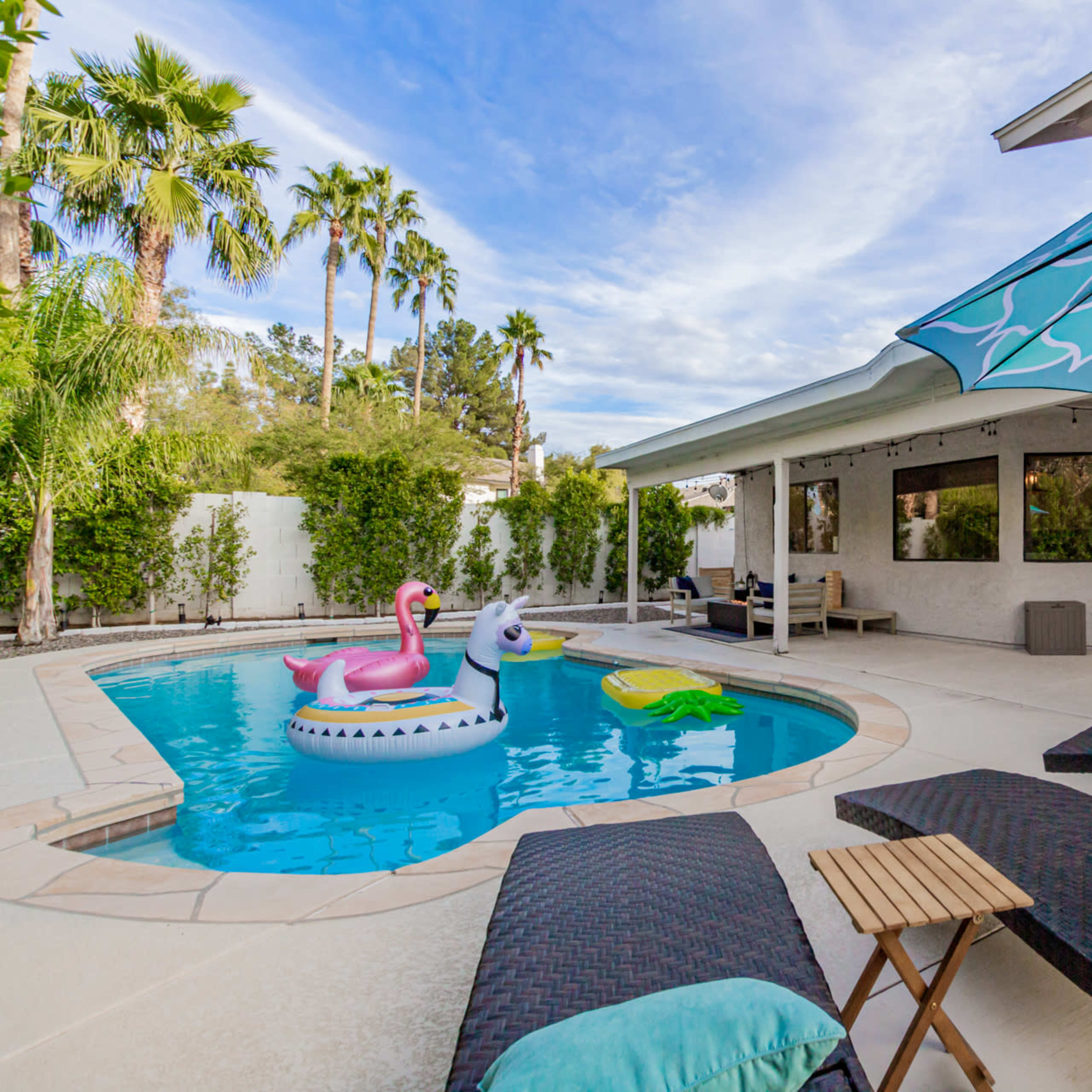 A backyard pool area featuring lounge chairs, a large umbrella, and inflatable pool toys in the water.