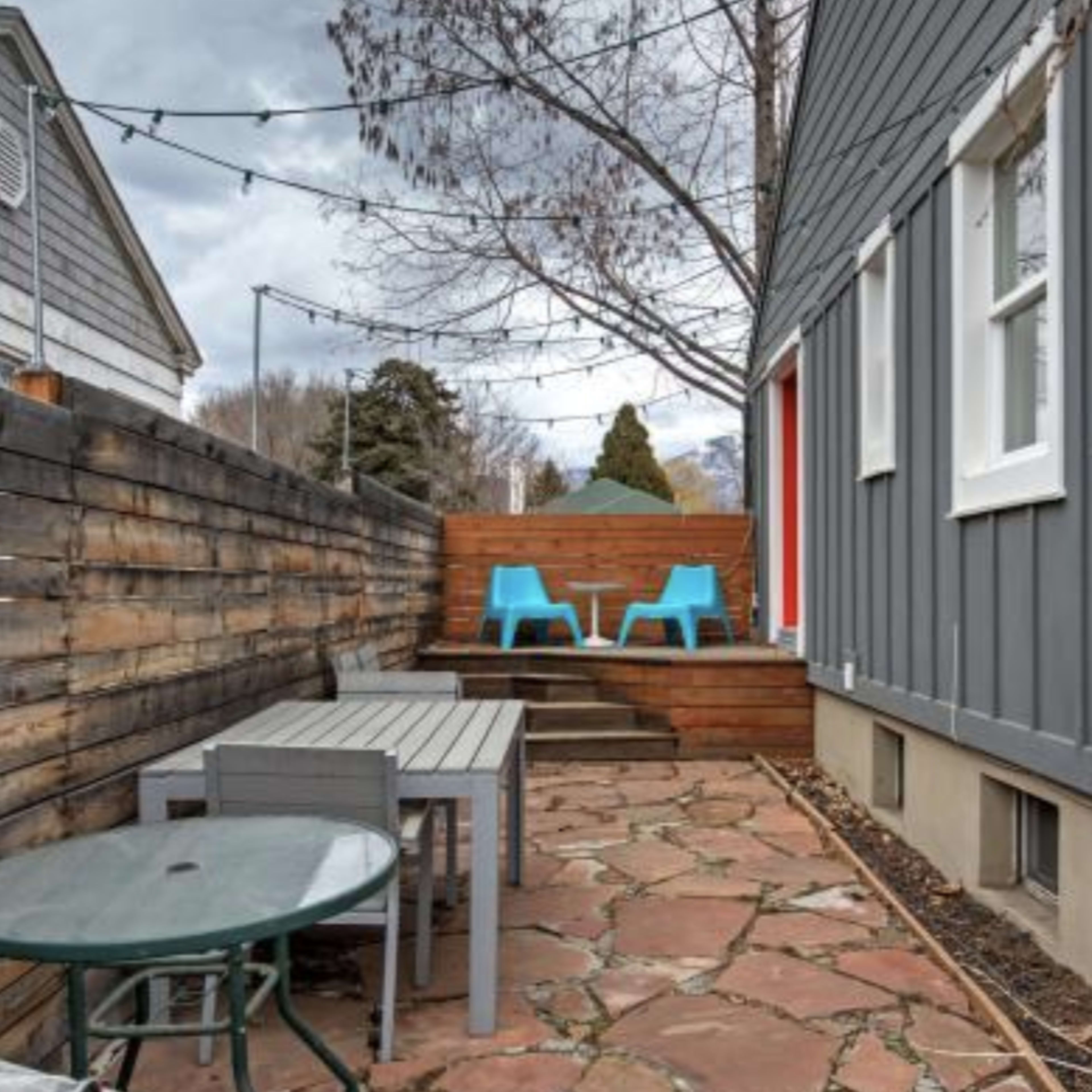 A small outdoor patio area with two blue chairs, a wooden step leading to a raised platform, and a stone pathway bordered by a wooden fence.