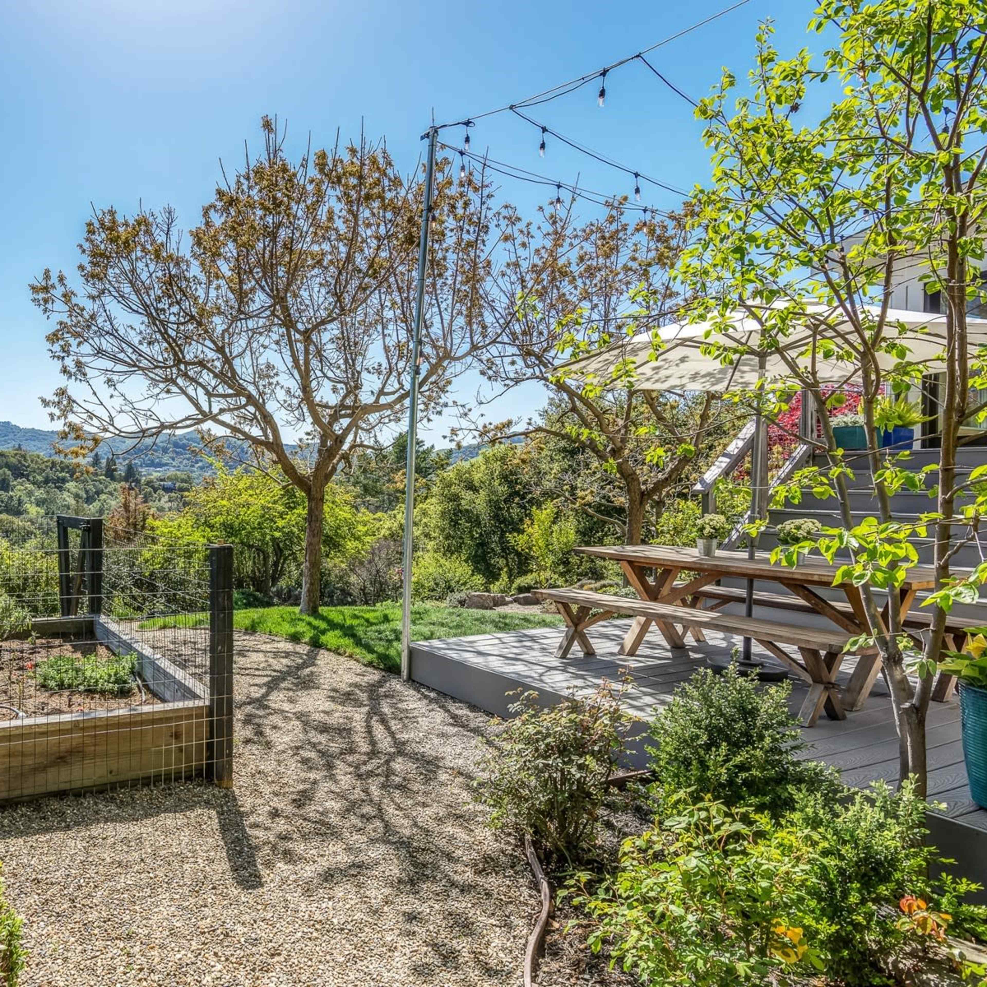 A landscaped backyard features a garden bed, a picnic table under a shade structure, and a view of rolling hills in the distance.
