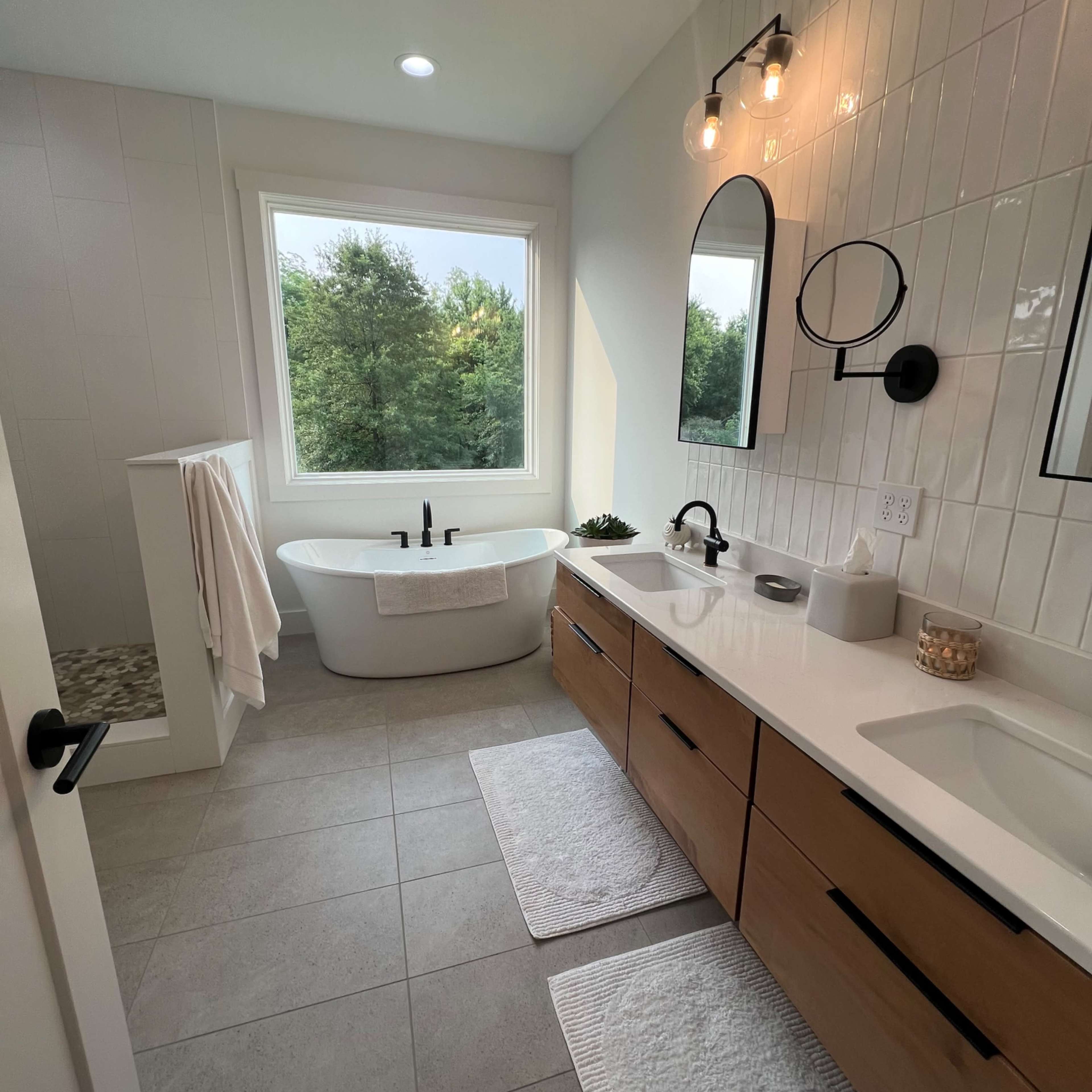 A modern bathroom featuring a freestanding bathtub, dual sink vanity with wooden cabinets, a large window overlooking greenery, and stylish lighting.