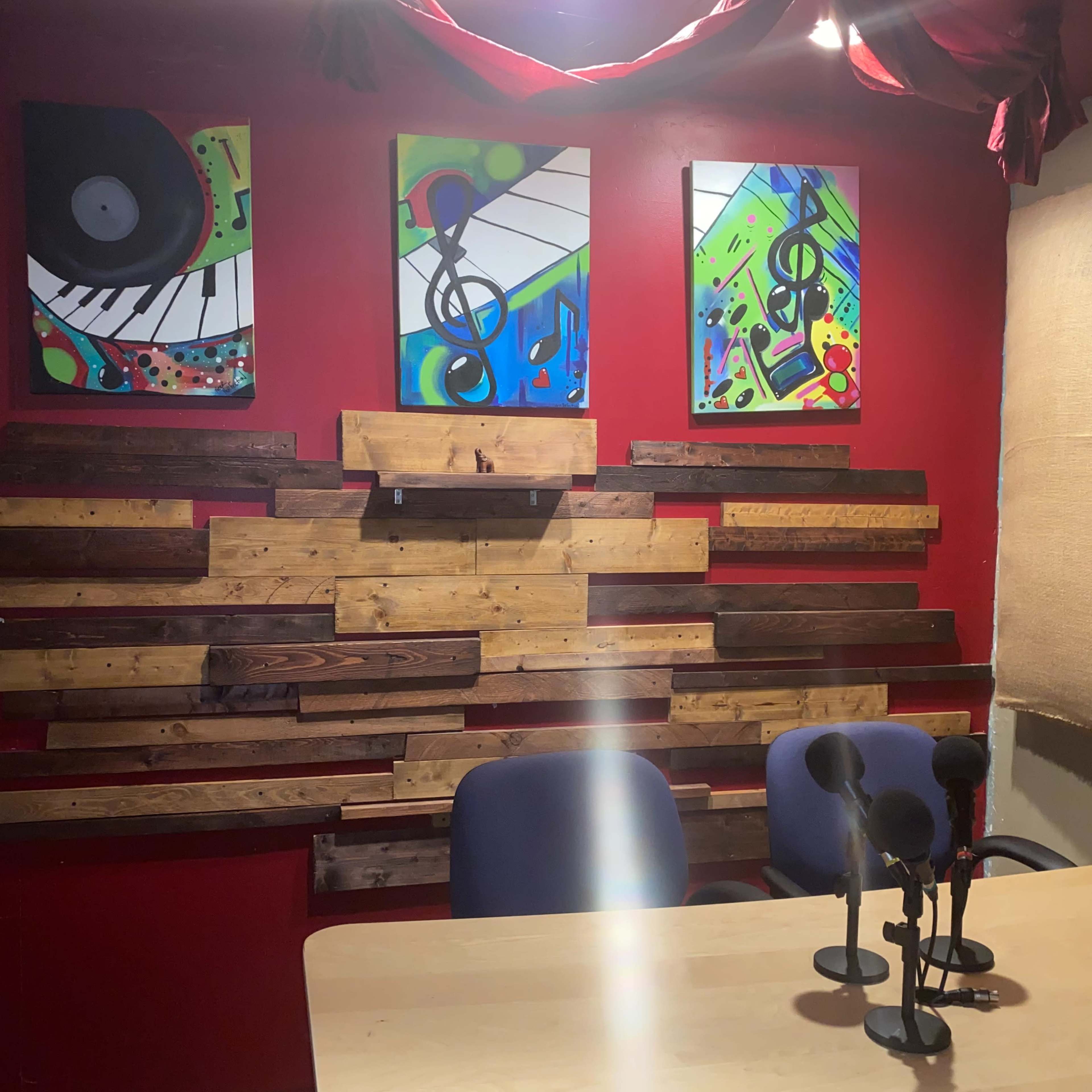 A small, brightly colored recording studio with a wooden slat wall, two blue chairs, and a table equipped with microphones.