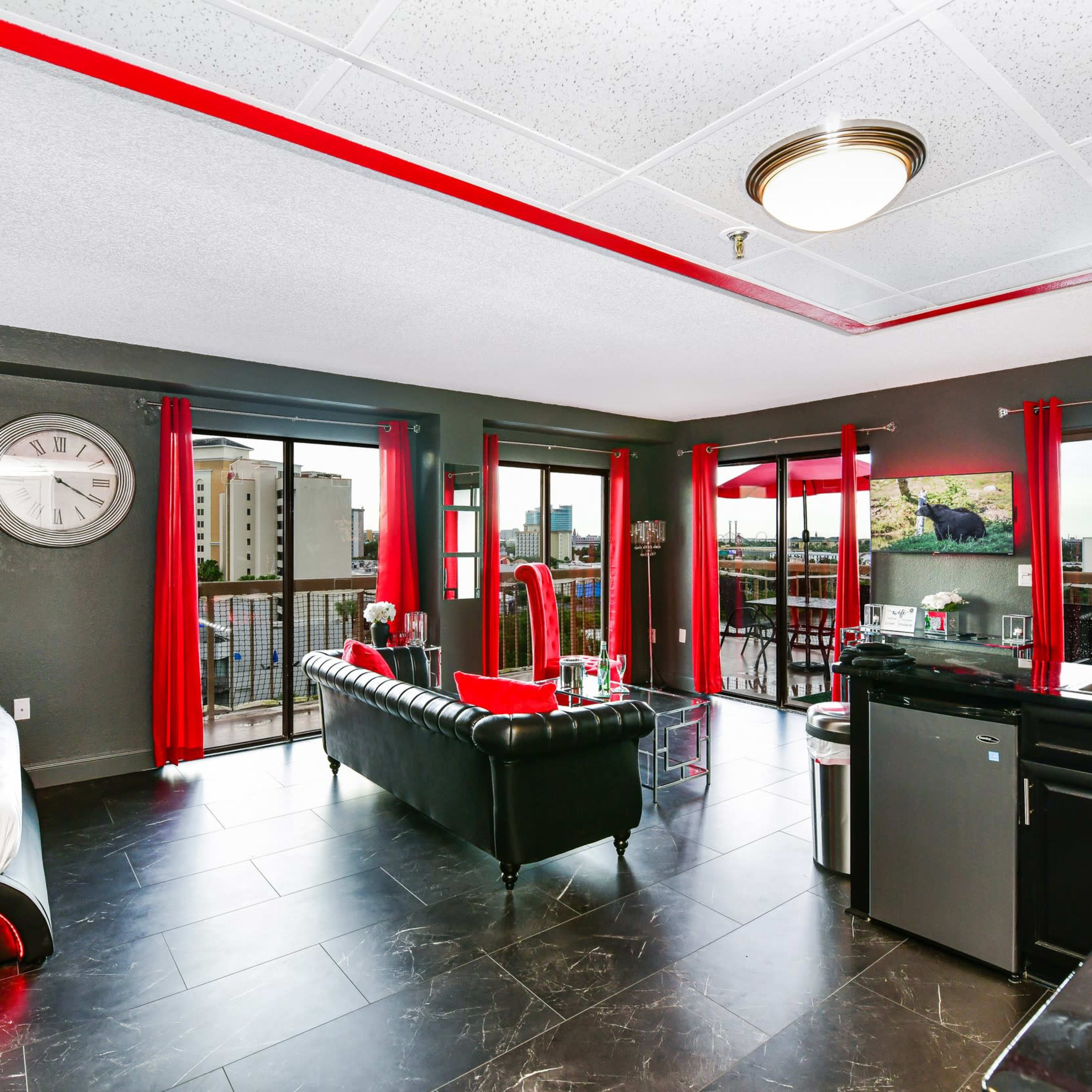 A modern apartment features a black and red color scheme with a living area, a bed, and large windows showing a city view.