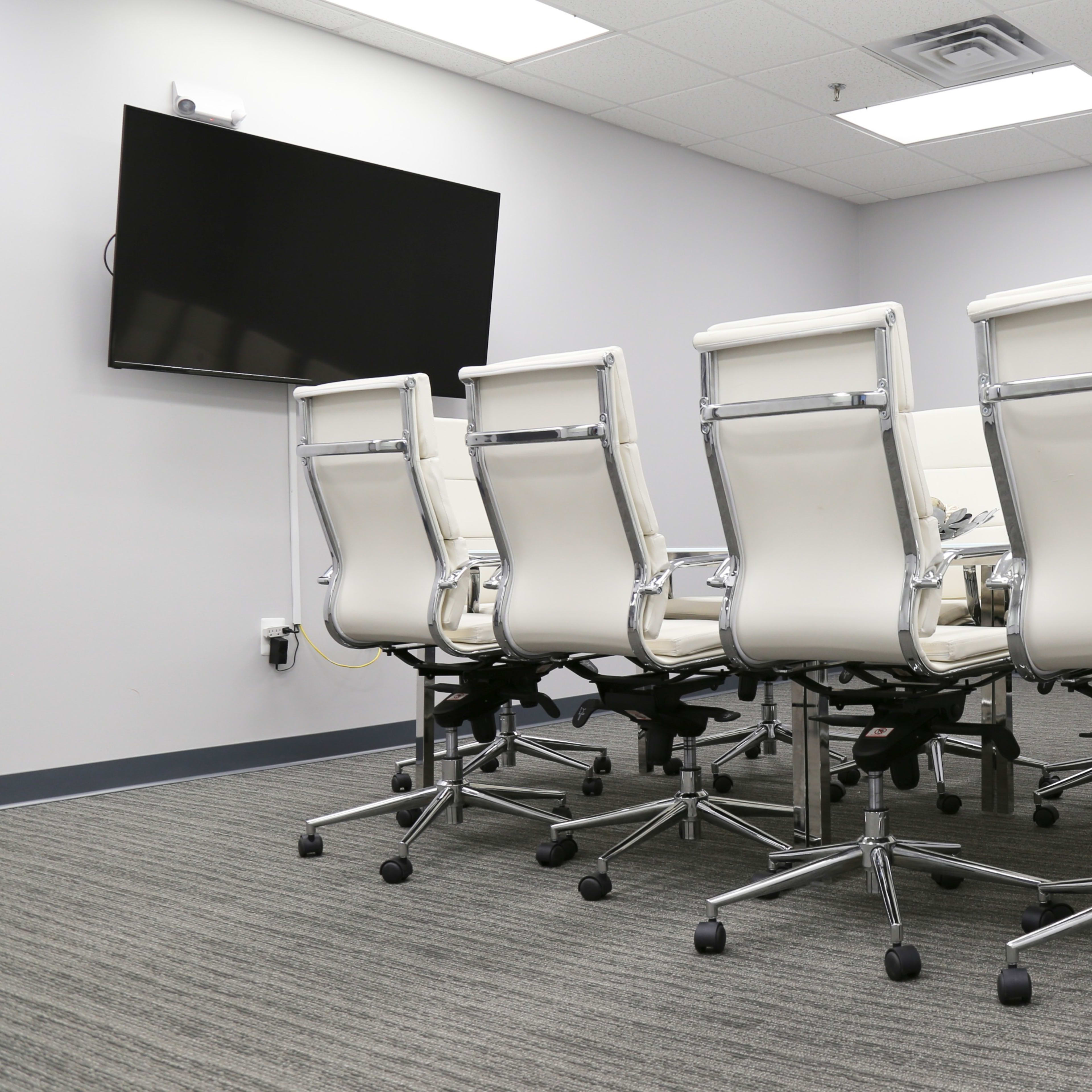 A conference room with a long table and several white office chairs arranged around it, and a wall-mounted TV at one end.