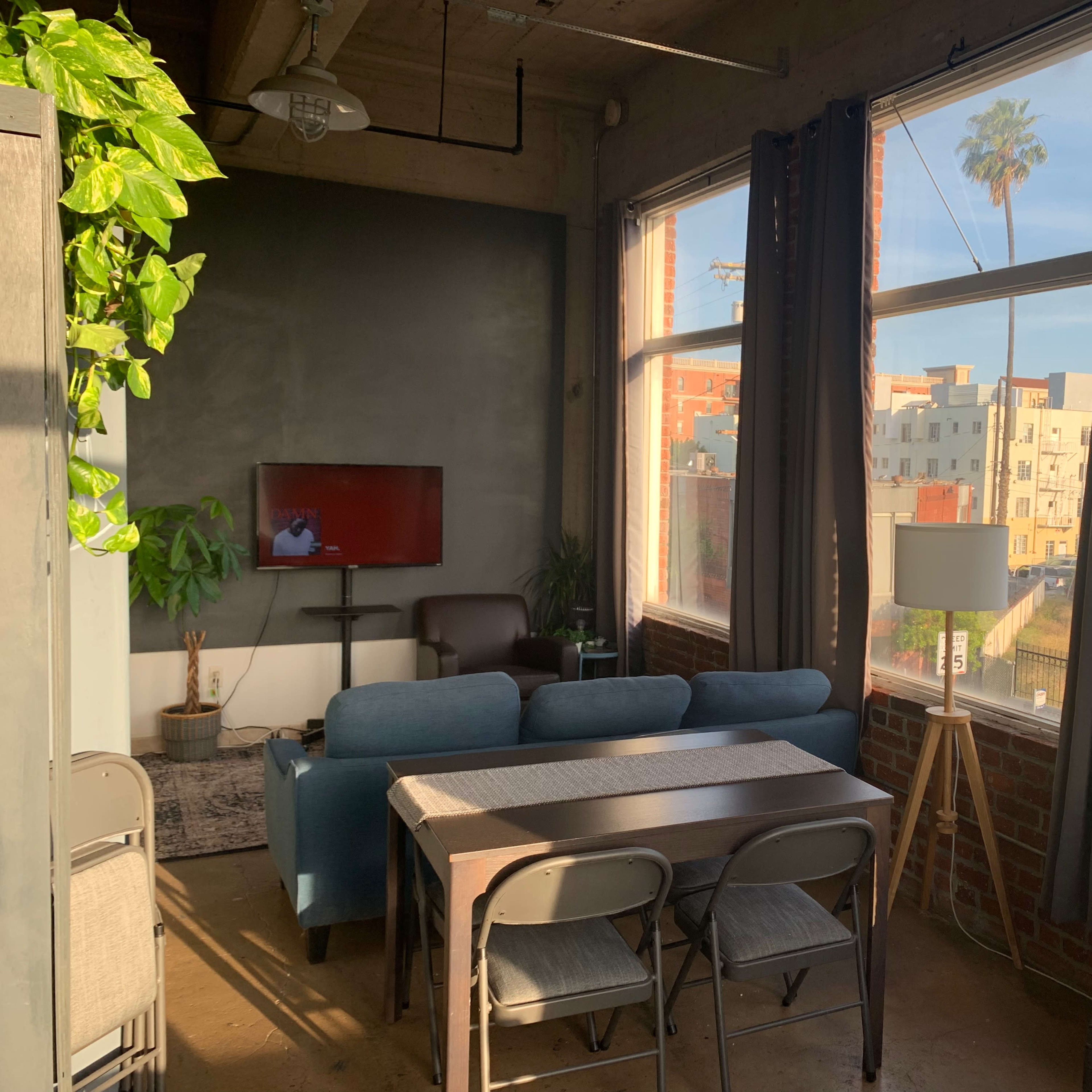 A living room with large windows allows natural light to illuminate a seating area, dining table, and television against a brick wall.
