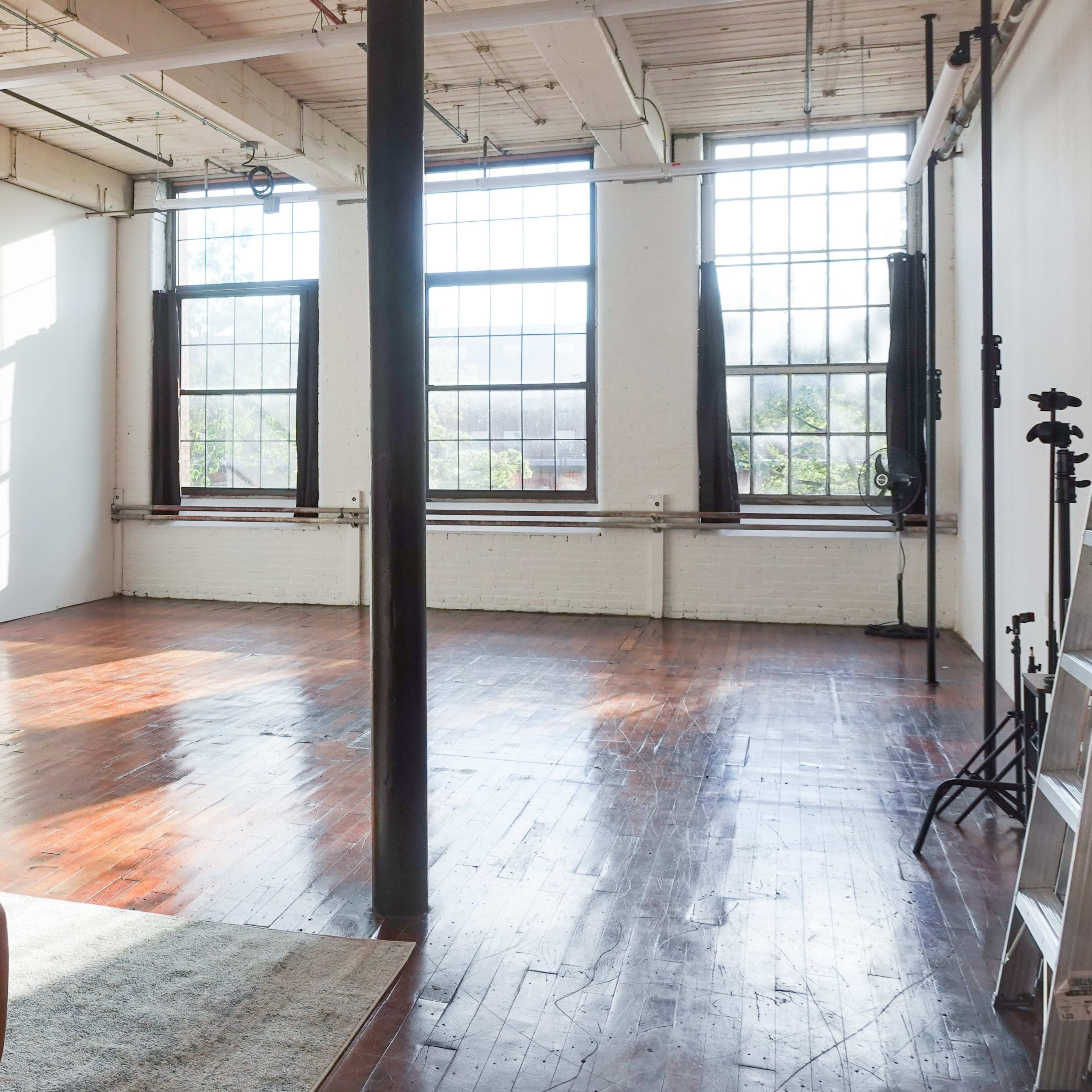 A spacious, empty studio with large windows that let in natural light, wooden floors, and minimal furnishings.