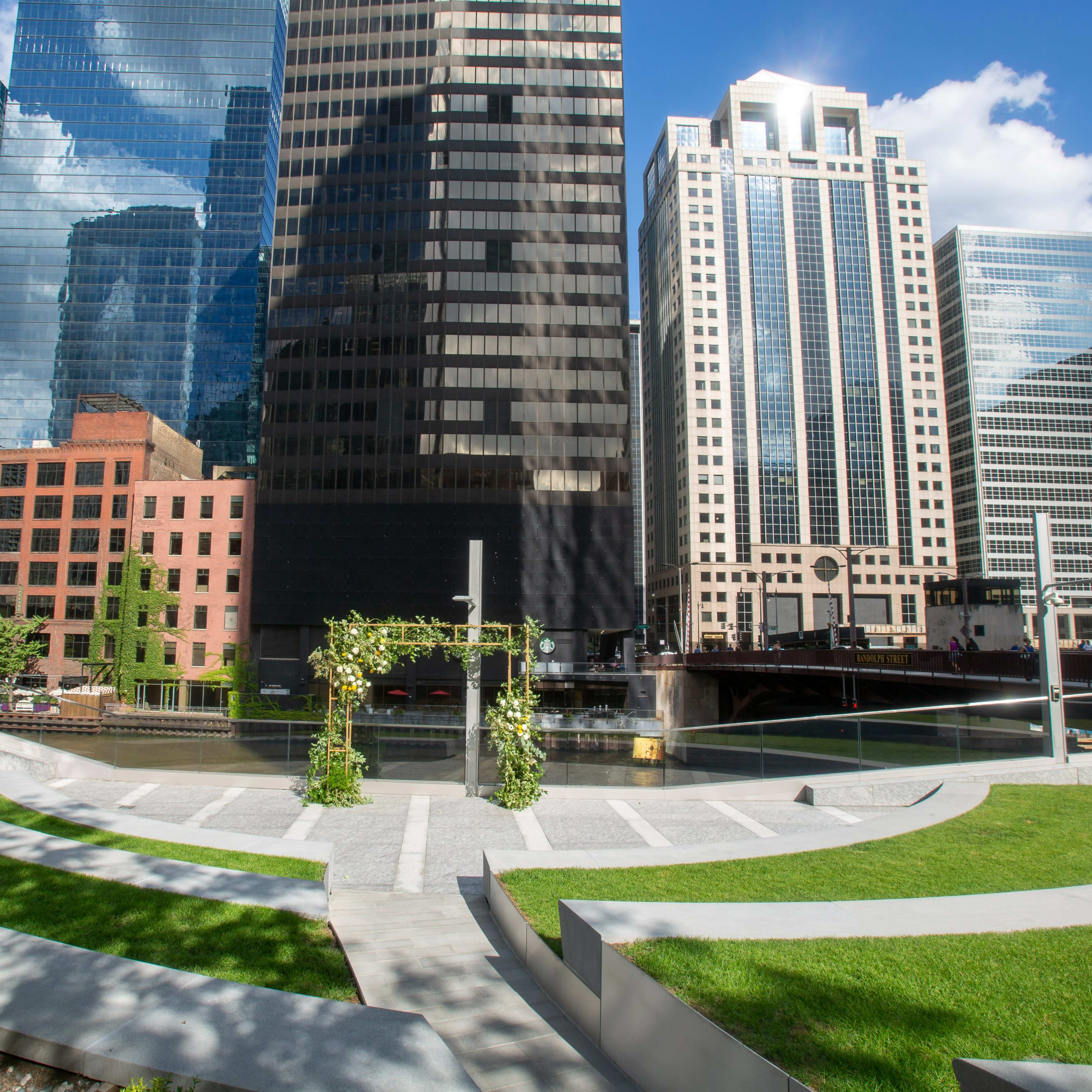 A modern urban park features curved seating areas and a floral arch set against a backdrop of tall skyscrapers and a river.