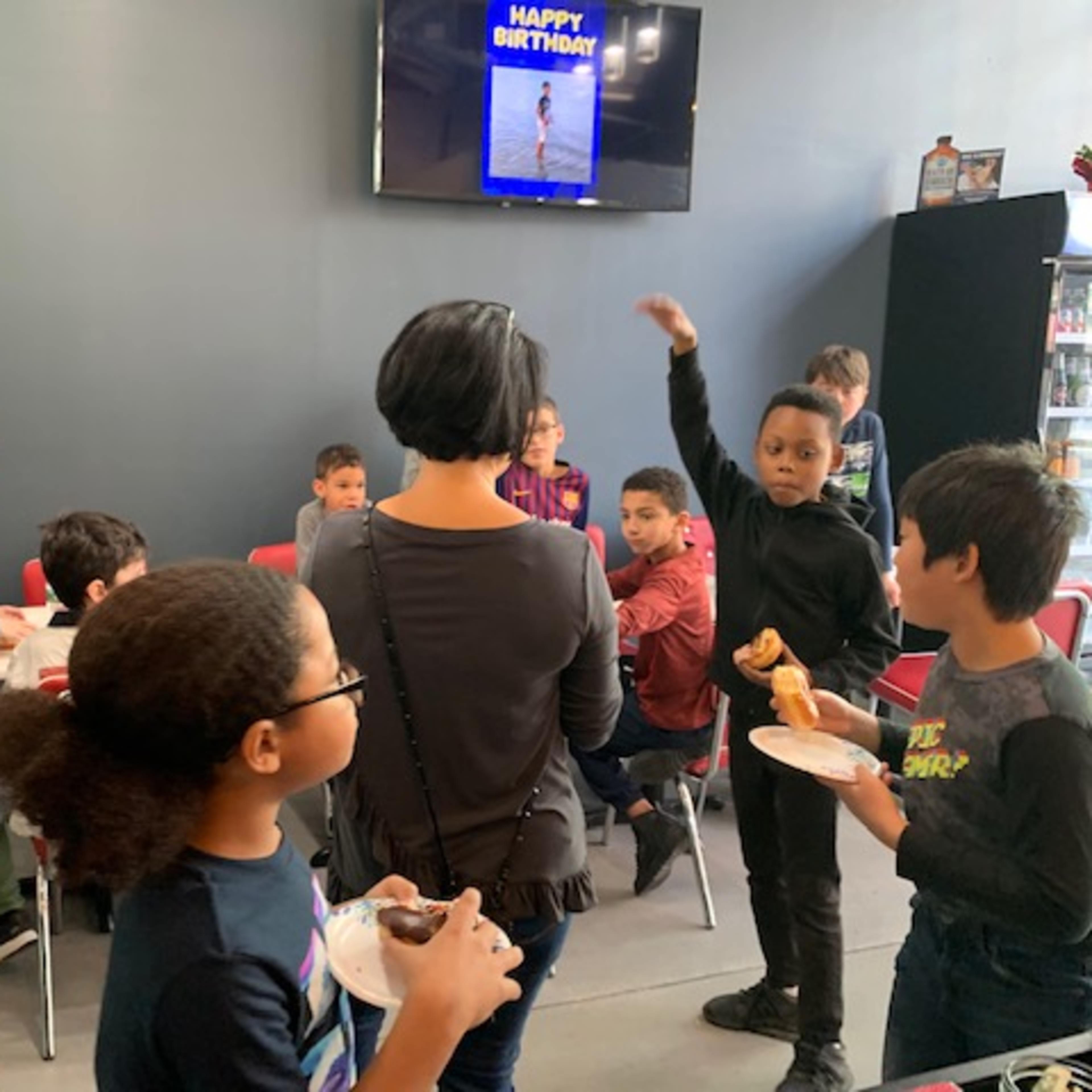 A group of children and adults are gathered in a party room, with some enjoying food and others interacting, while a digital sign displays "HAPPY BIRTHDAY" on the wall.