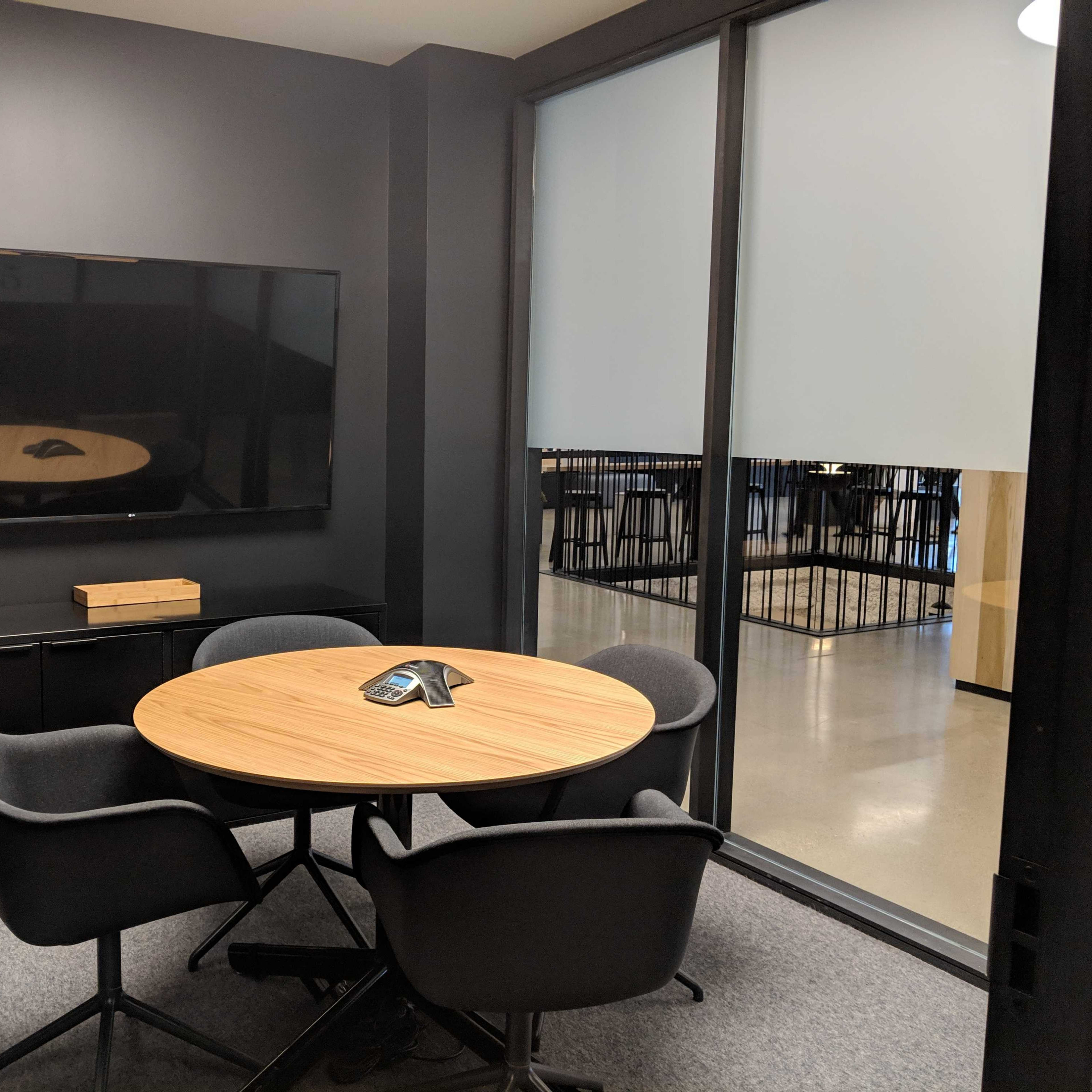A small conference room features a round wooden table surrounded by four gray chairs, with a large screen mounted on the wall.