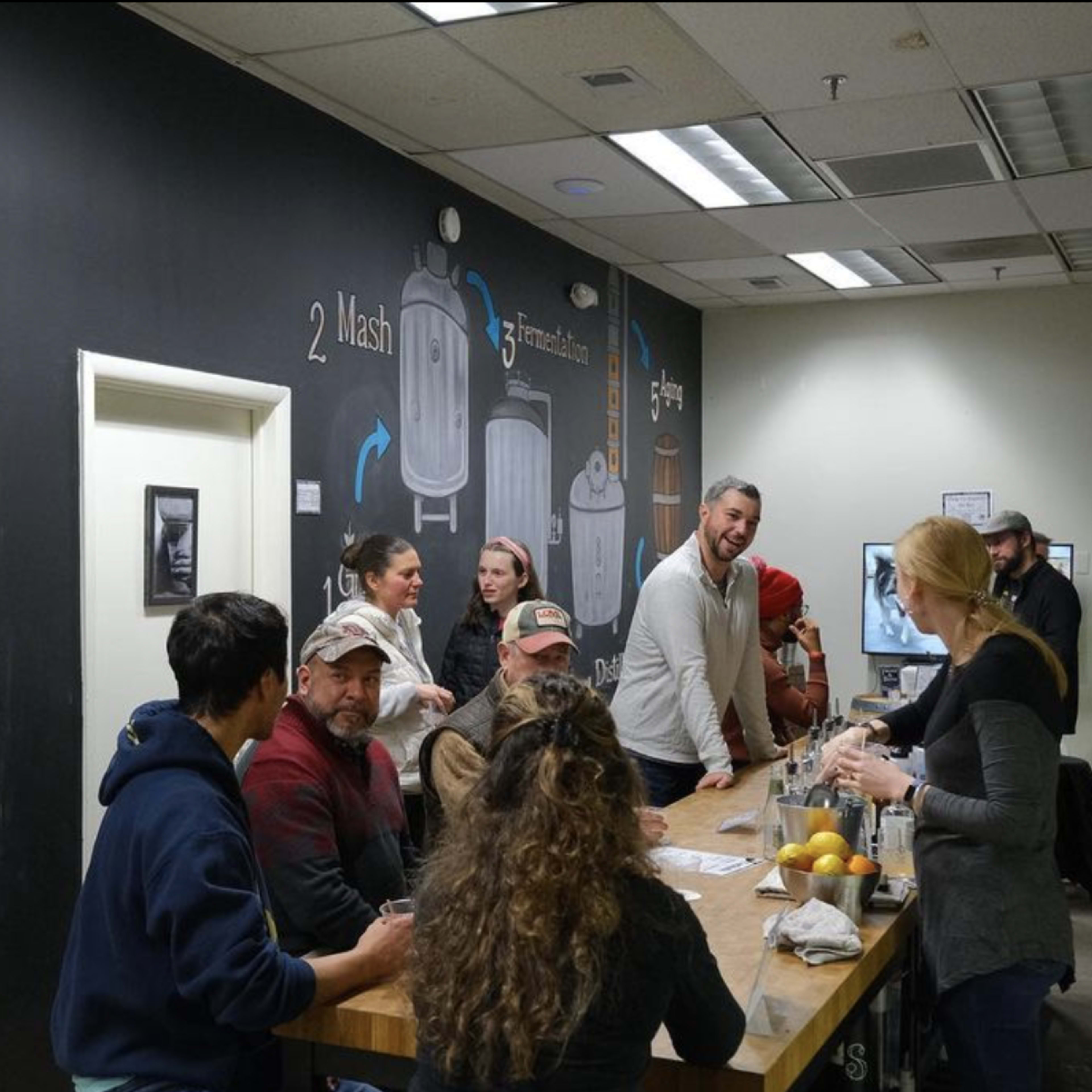 A group of people is gathered around a wooden table in a brewery, engaging with staff and tasting various beverages in a room decorated with chalkboard illustrations of brewing equipment.