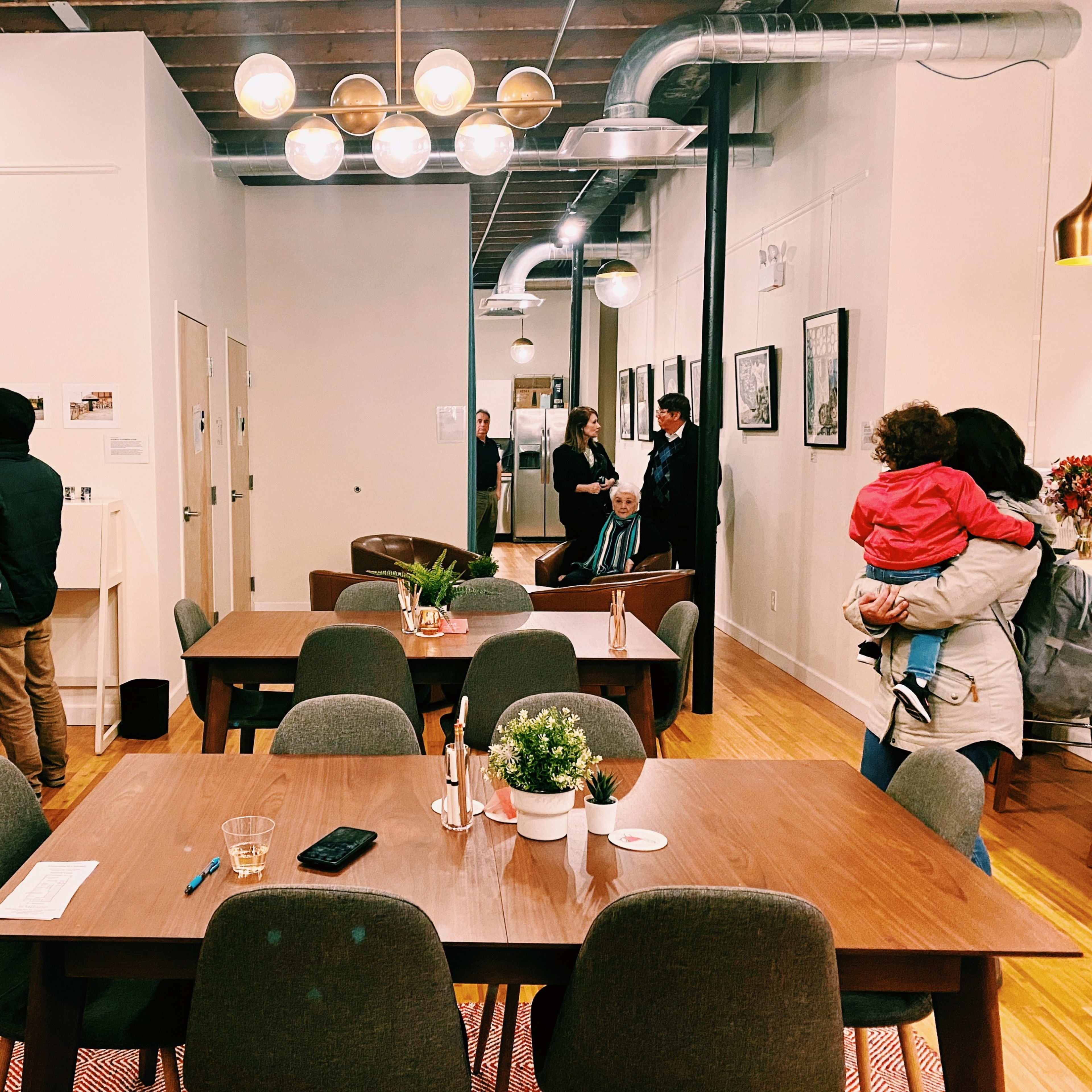 A spacious interior with wooden tables and chairs, where several people are engaged in various activities, including browsing a wall display and conversing.