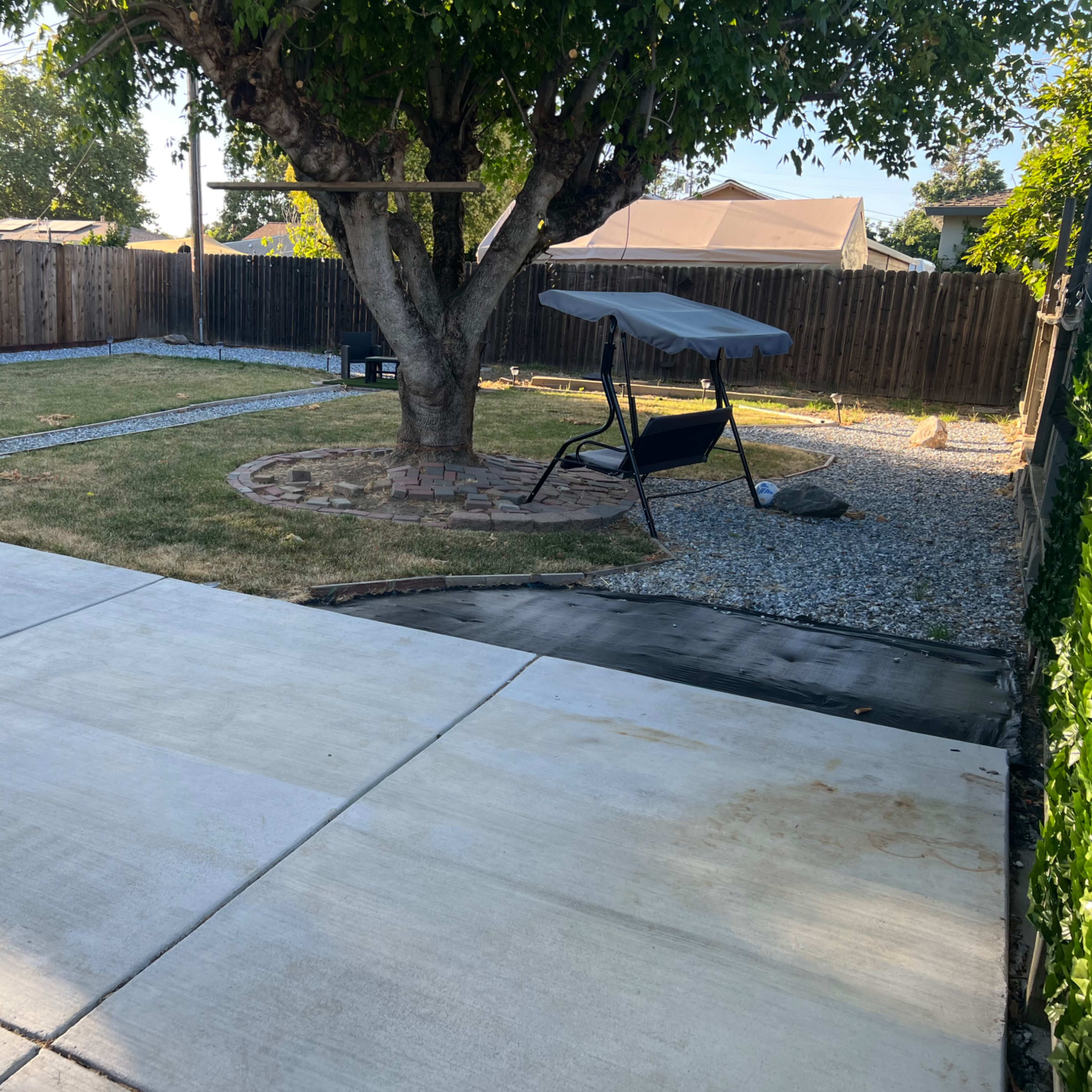 A backyard with a large tree, a gravel circle, a swing seat, and a lawn area bordered by a wooden fence.