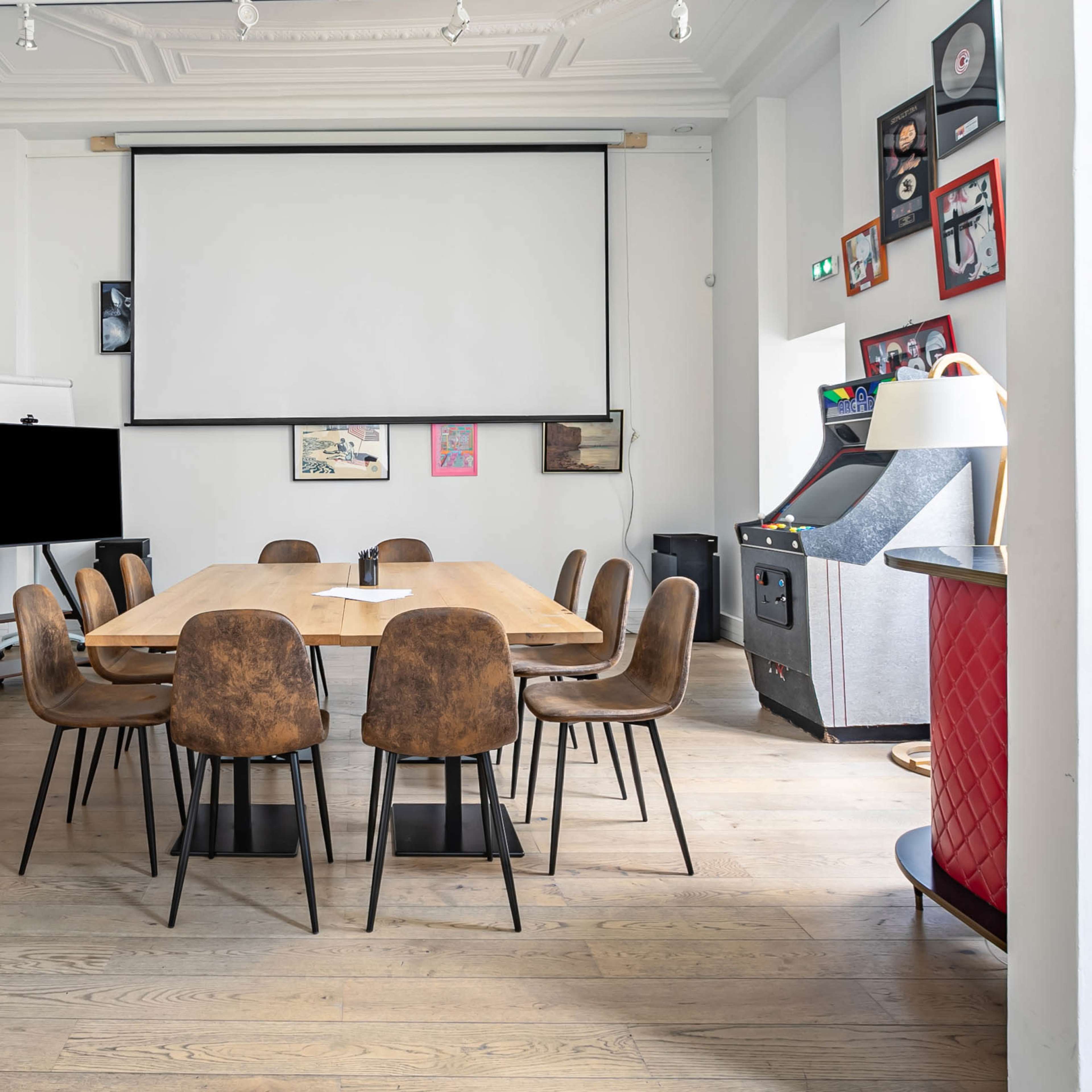 A conference room features a long wooden table surrounded by chairs, with shelves of books and a projector screen on the wall.
