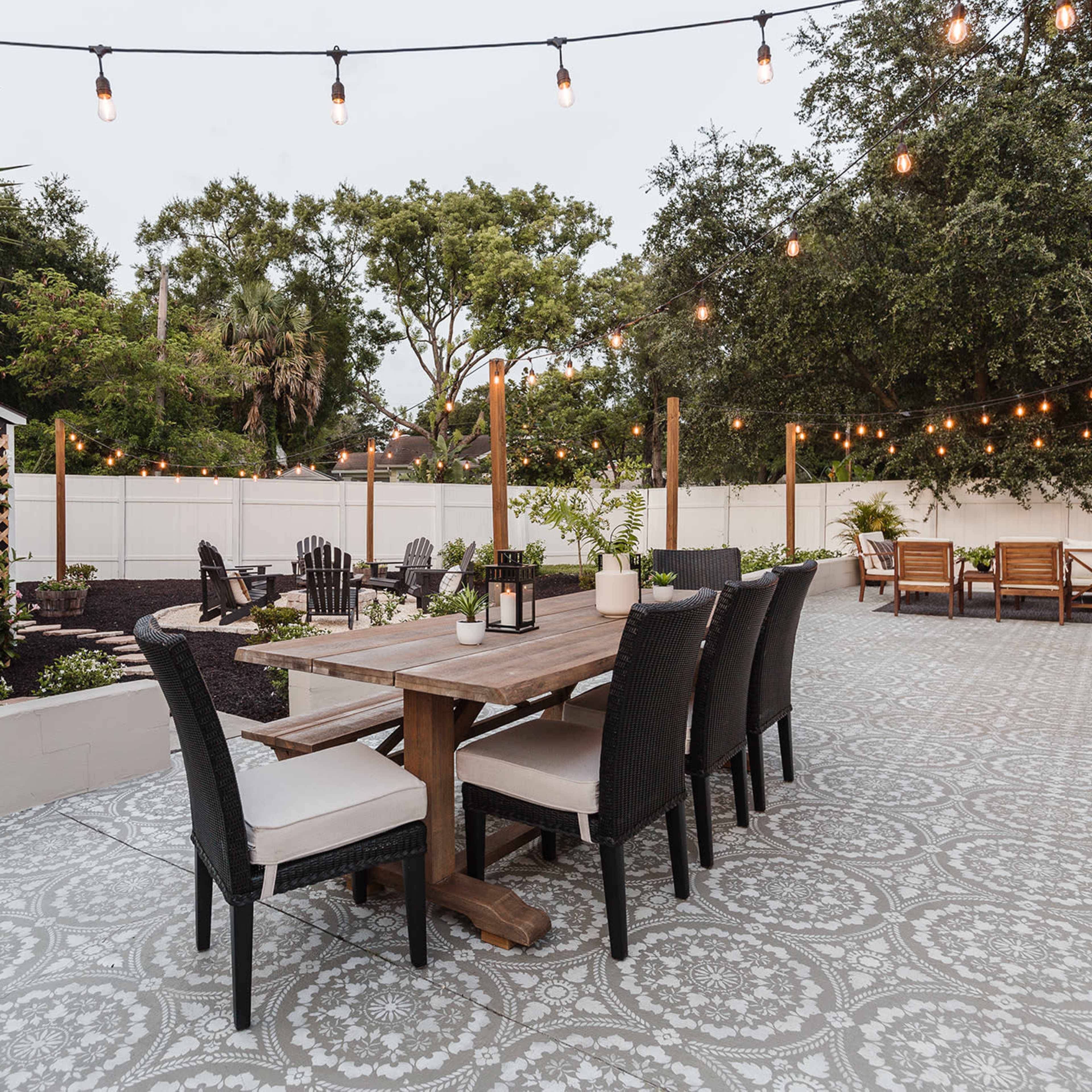 A spacious outdoor patio features a large wooden dining table surrounded by black chairs, with hanging string lights and greenery in the background.
