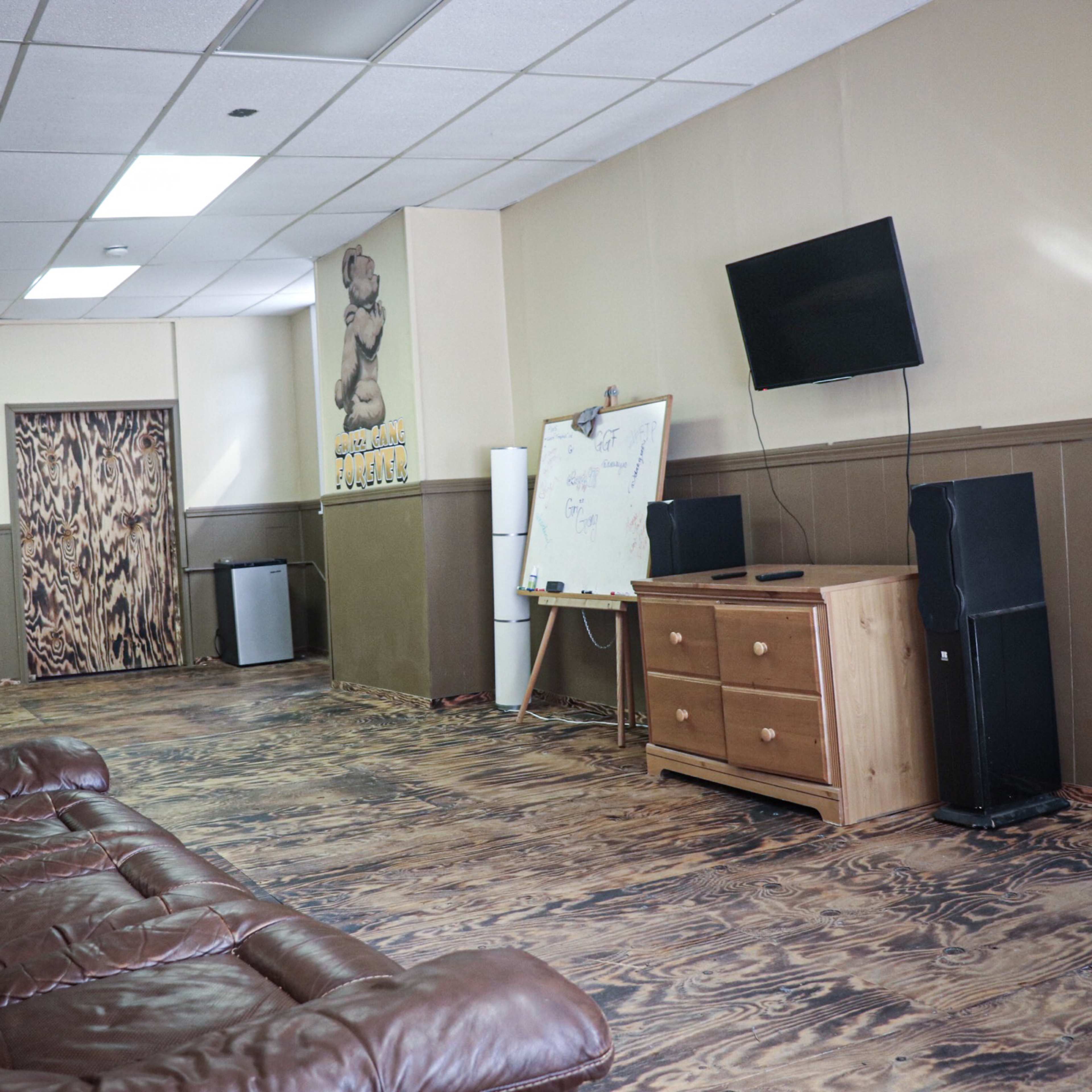 A long, sparsely furnished room with a brown leather sofa, a wooden dresser, a television mounted on the wall, and a whiteboard on an easel.