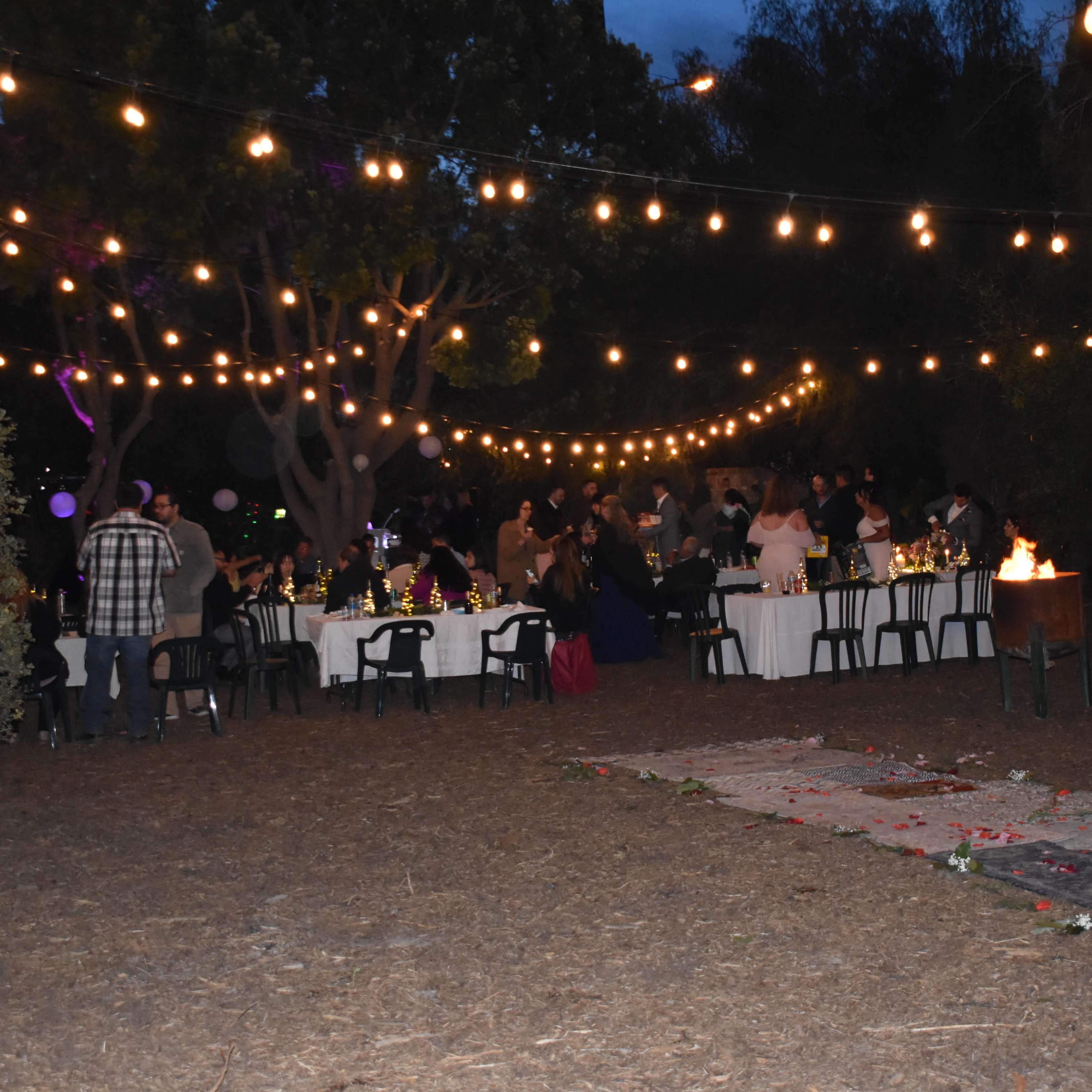 A group of people gathers around elegantly set tables under string lights in an outdoor setting, with a fire pit nearby.