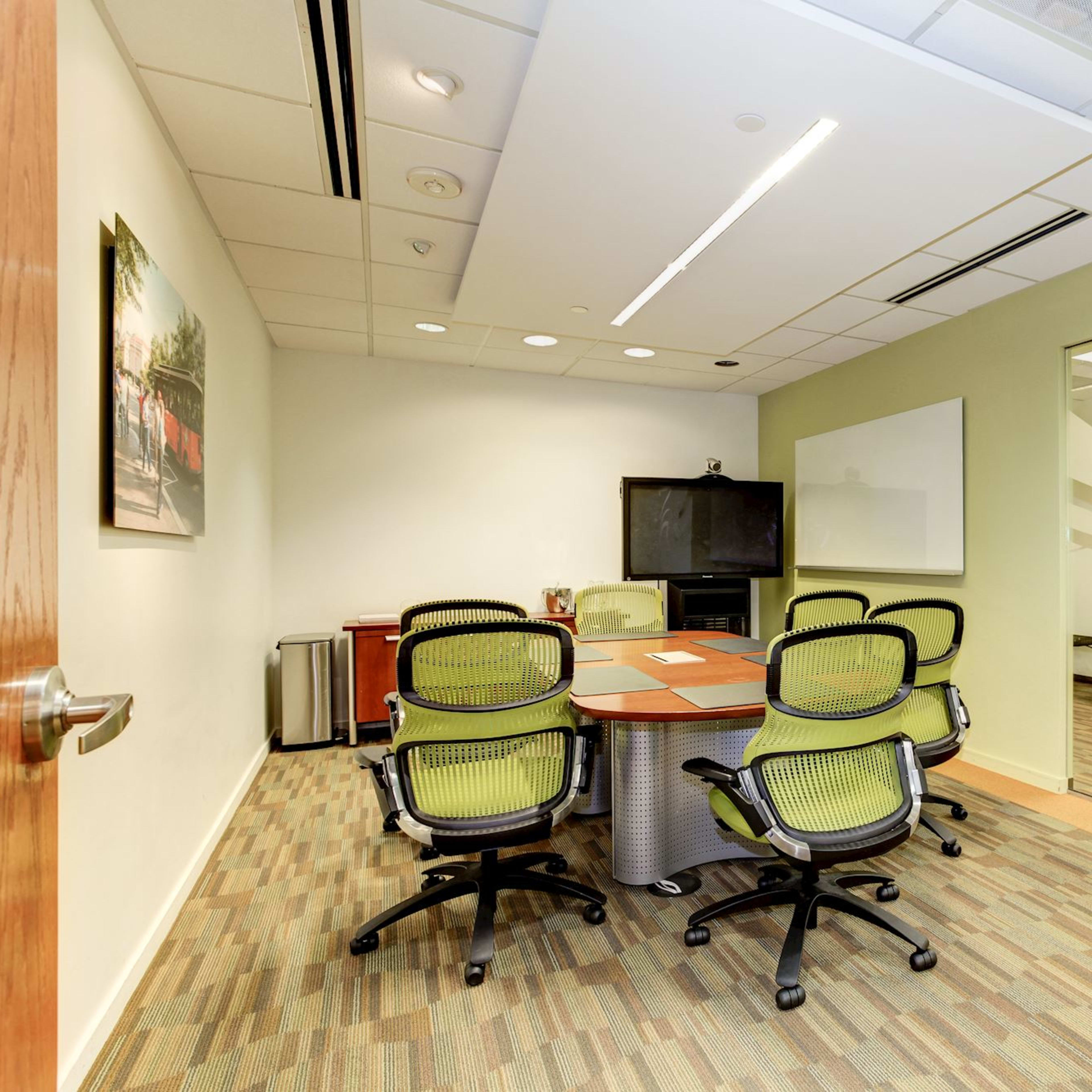 A modern conference room features a round table surrounded by green office chairs, with a television and whiteboard visible on the walls.
