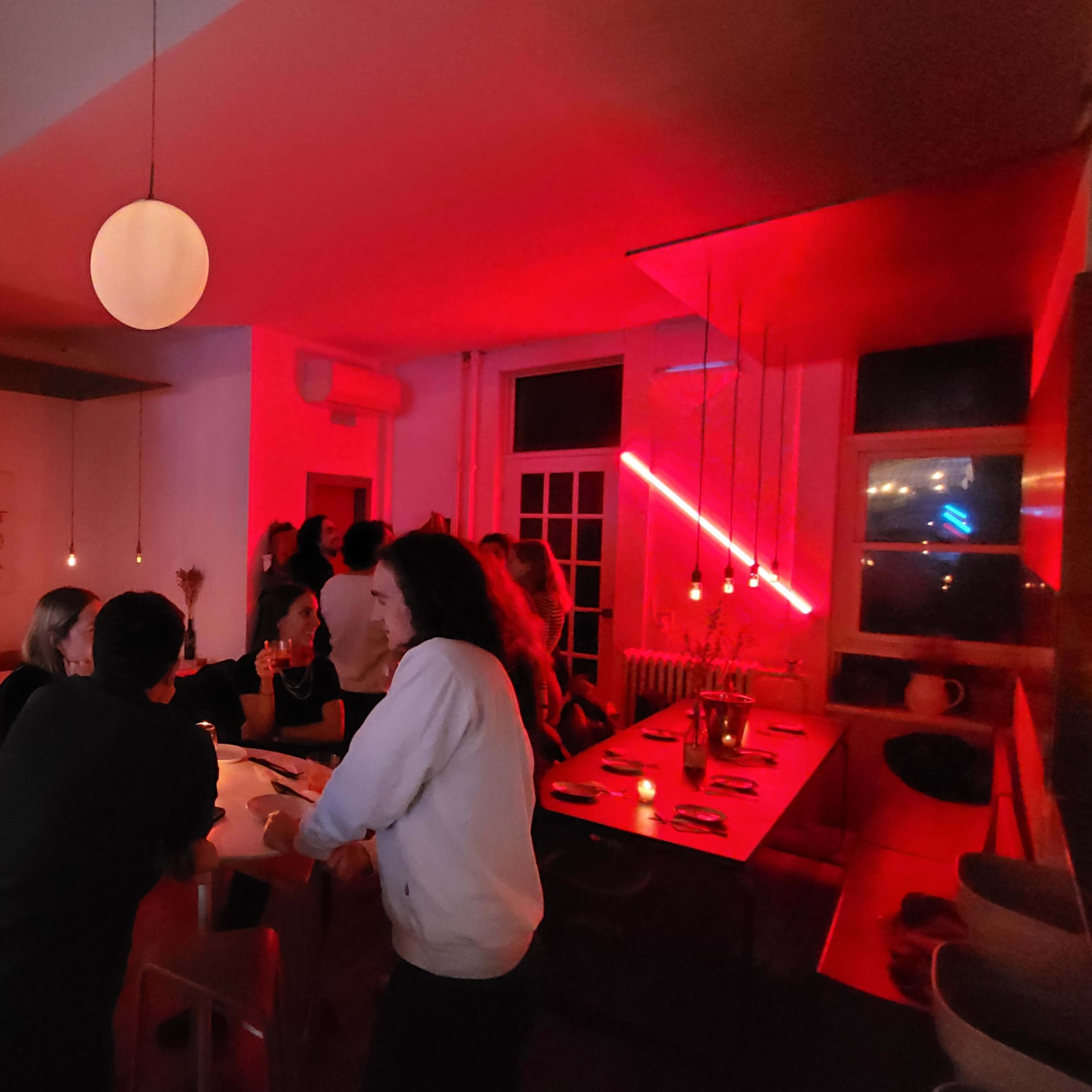The scene depicts a lively restaurant with patrons gathered at the bar and tables, illuminated by soft red lighting and pendant lamps.