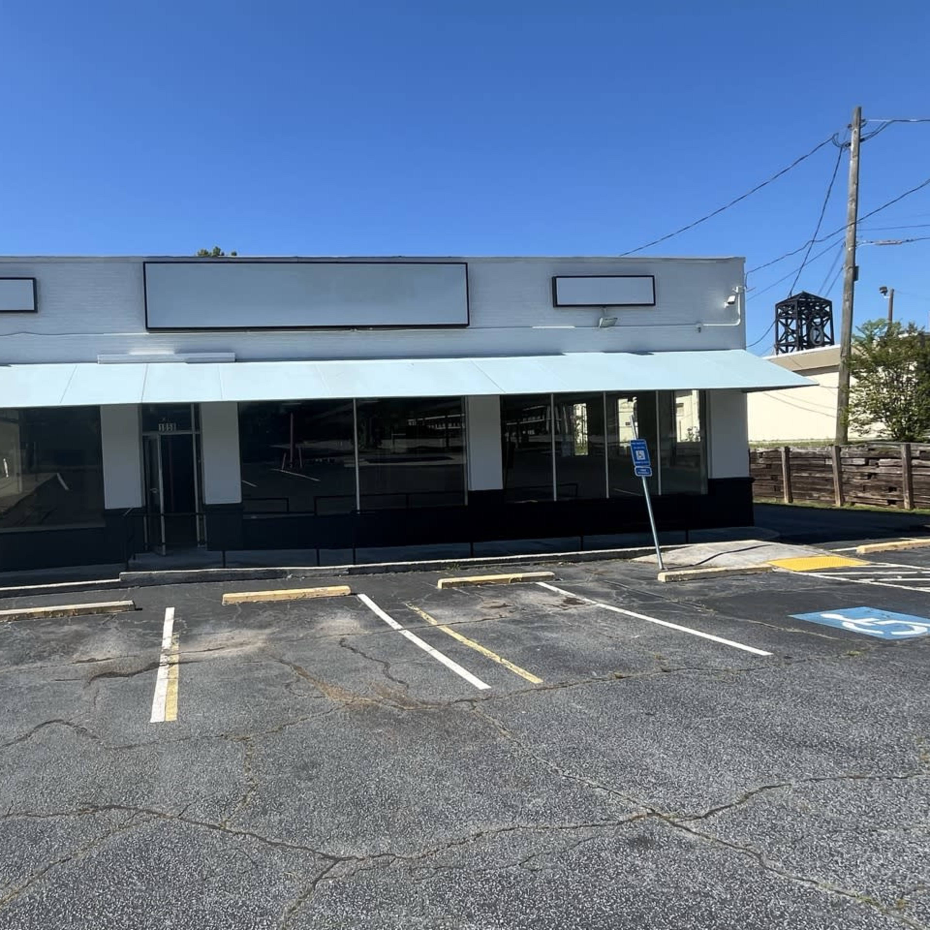A vacant commercial building with a large awning and multiple windows sits in an empty parking lot under a clear blue sky.