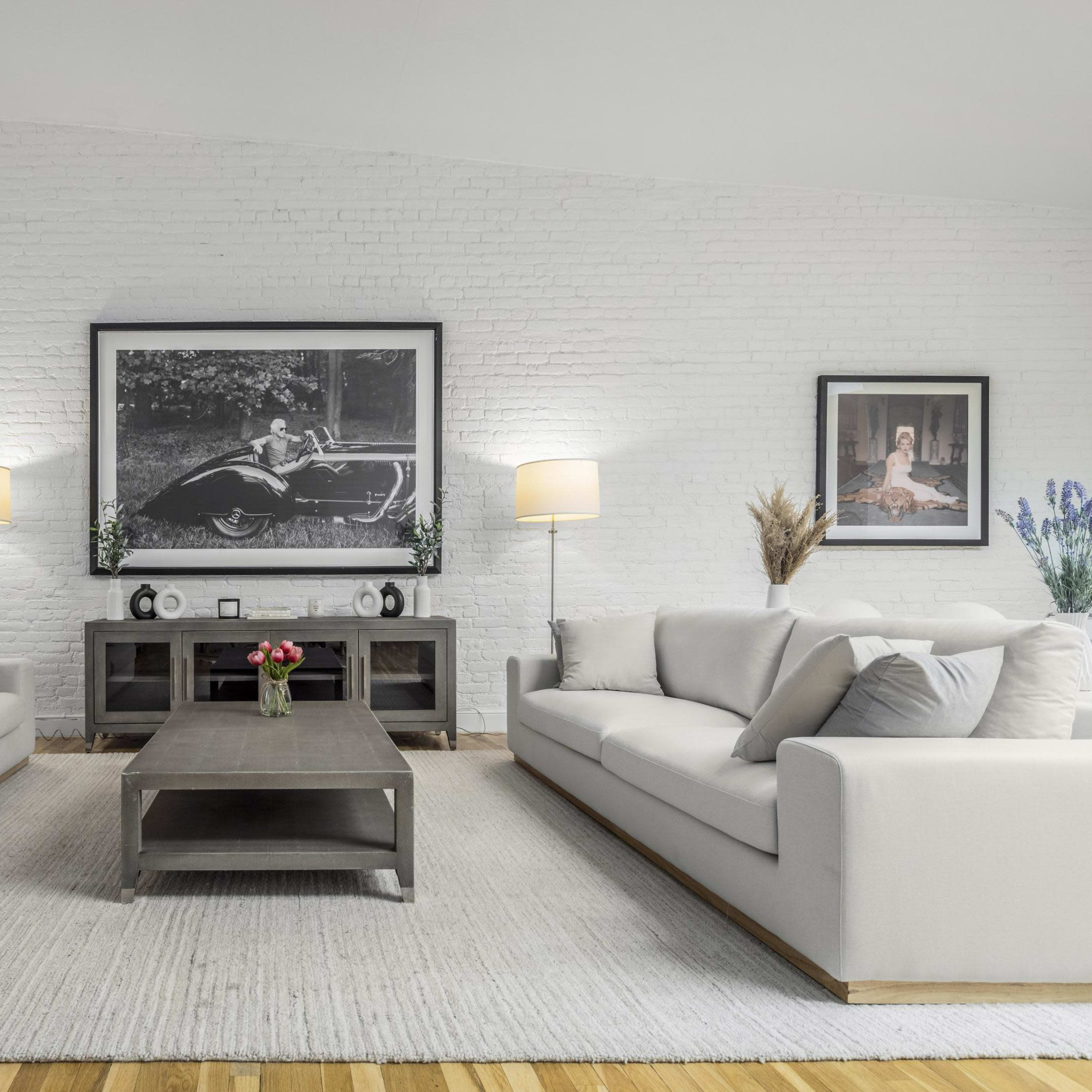 A modern living room with two light-colored sofas facing a central coffee table, against a backdrop of a white brick wall adorned with framed pictures and two floor lamps.