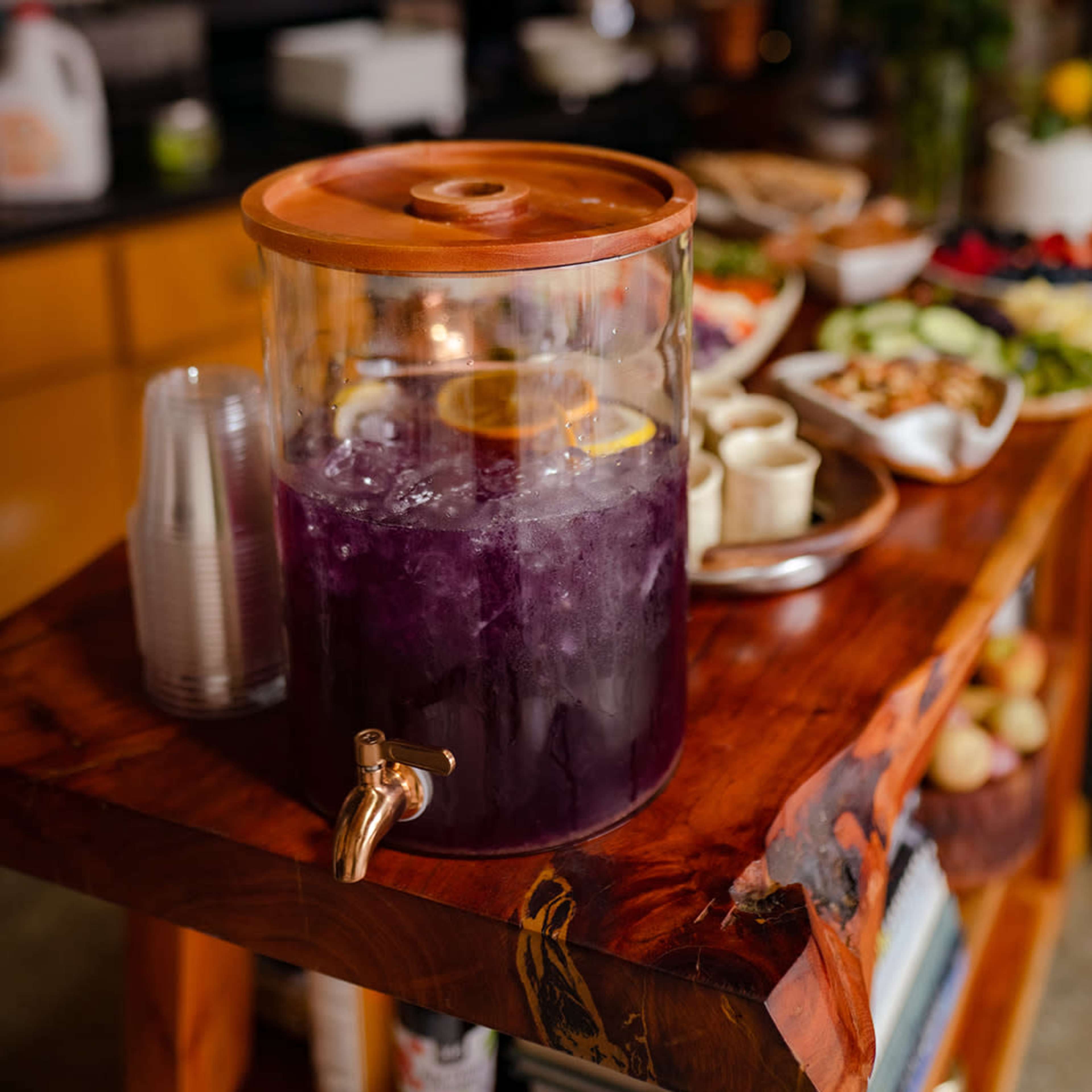 A glass beverage dispenser filled with purple drink and lemon slices sits on a wooden table alongside a spread of appetizers and flowers.