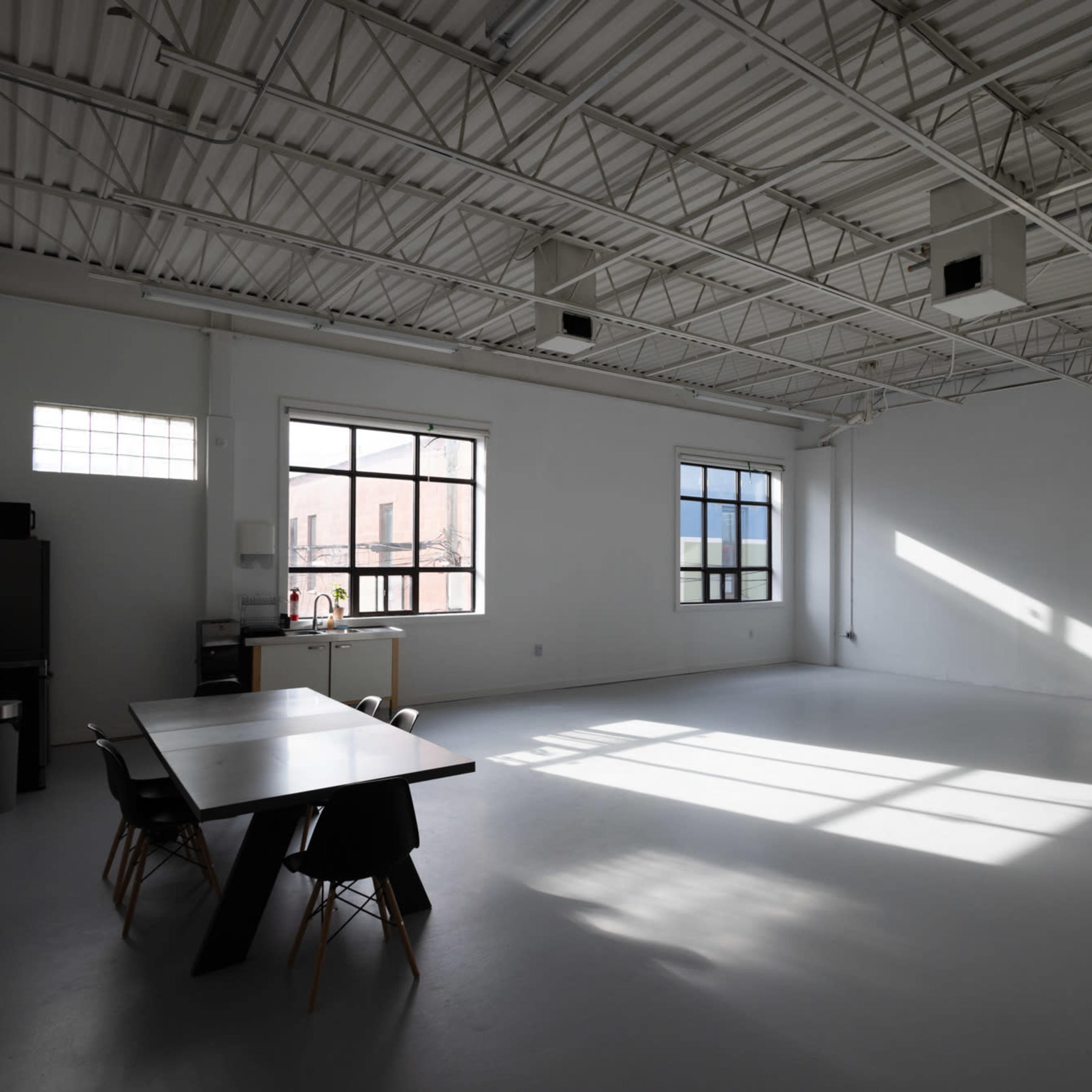 A large, empty room with white walls, a long table, and several chairs, illuminated by sunlight streaming through the windows.