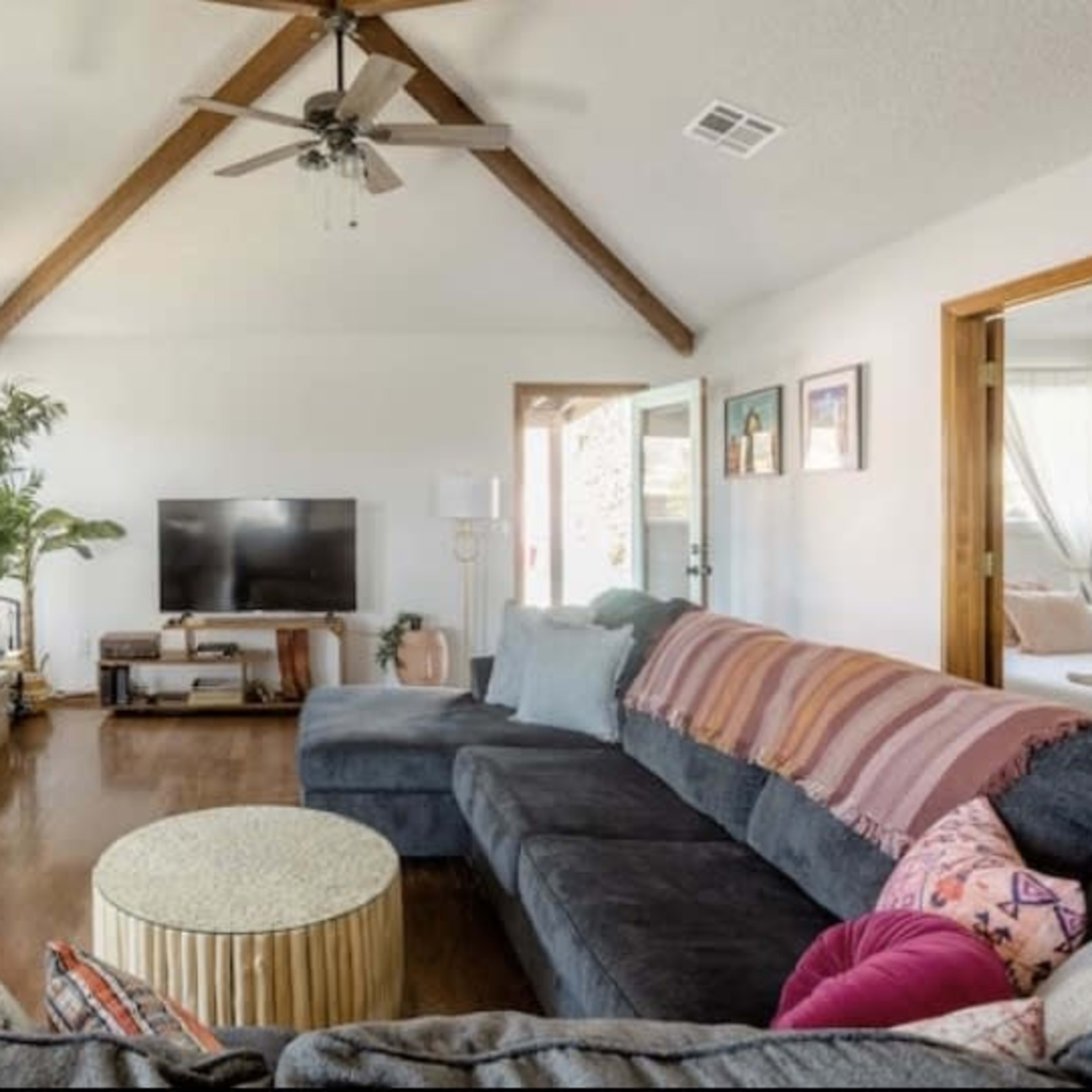 The living room features a stone fireplace, a large sectional sofa with colorful throw pillows, and a round coffee table, all set under exposed wooden beams.