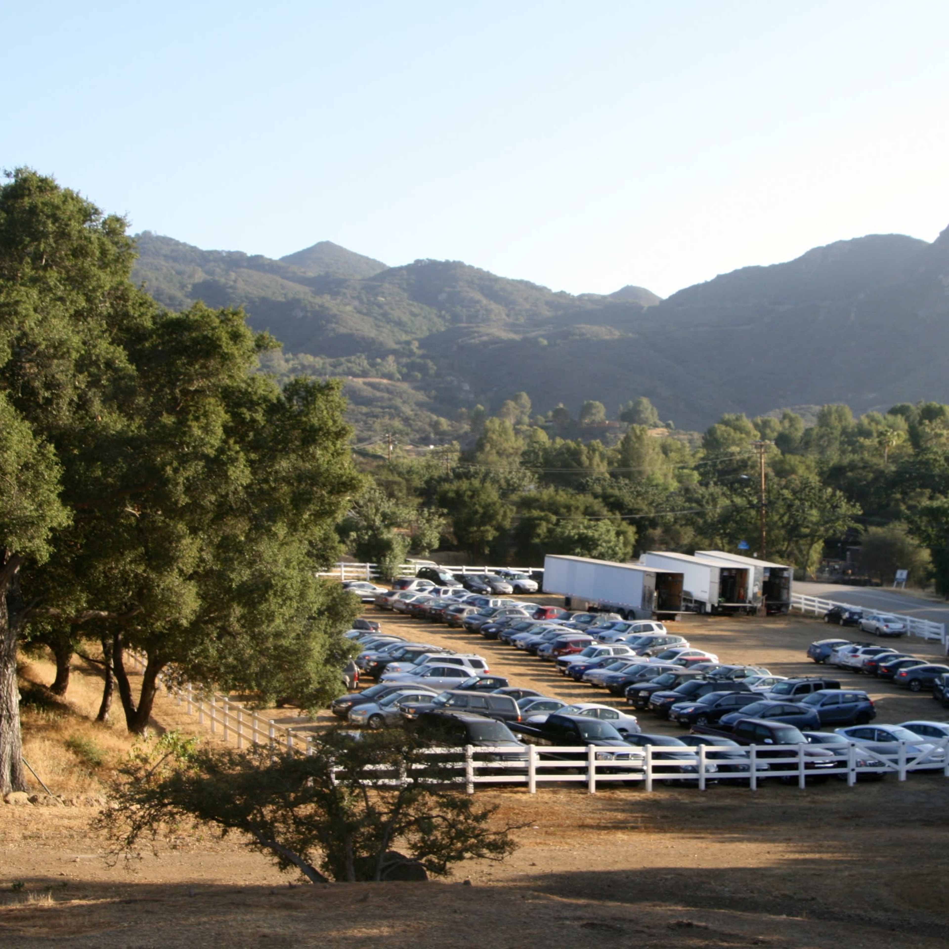 A large parking lot filled with cars is situated in a rural landscape surrounded by trees and mountains.
