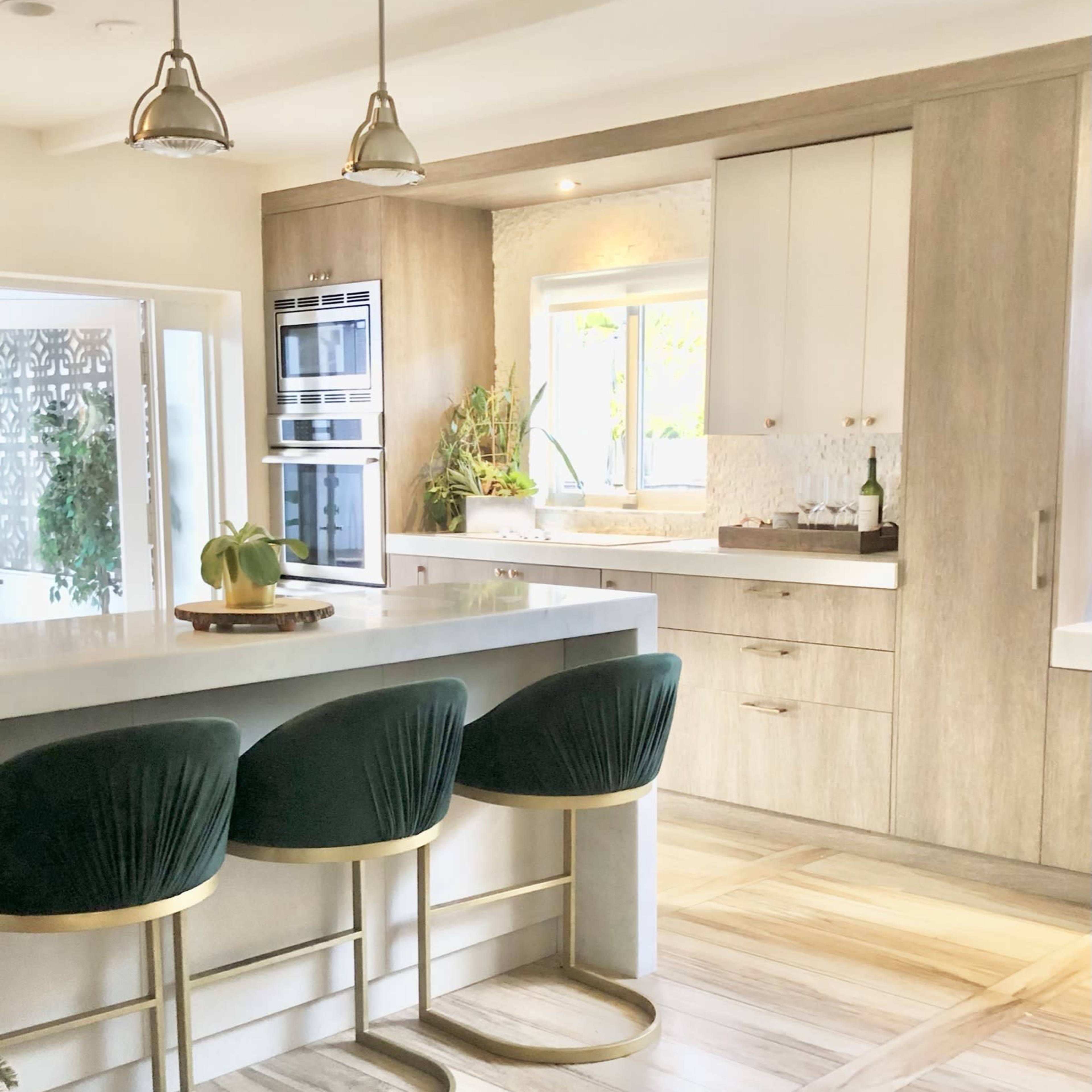 A modern kitchen featuring a light-colored countertop with three green bar stools and sleek cabinetry, illuminated by pendant lights.