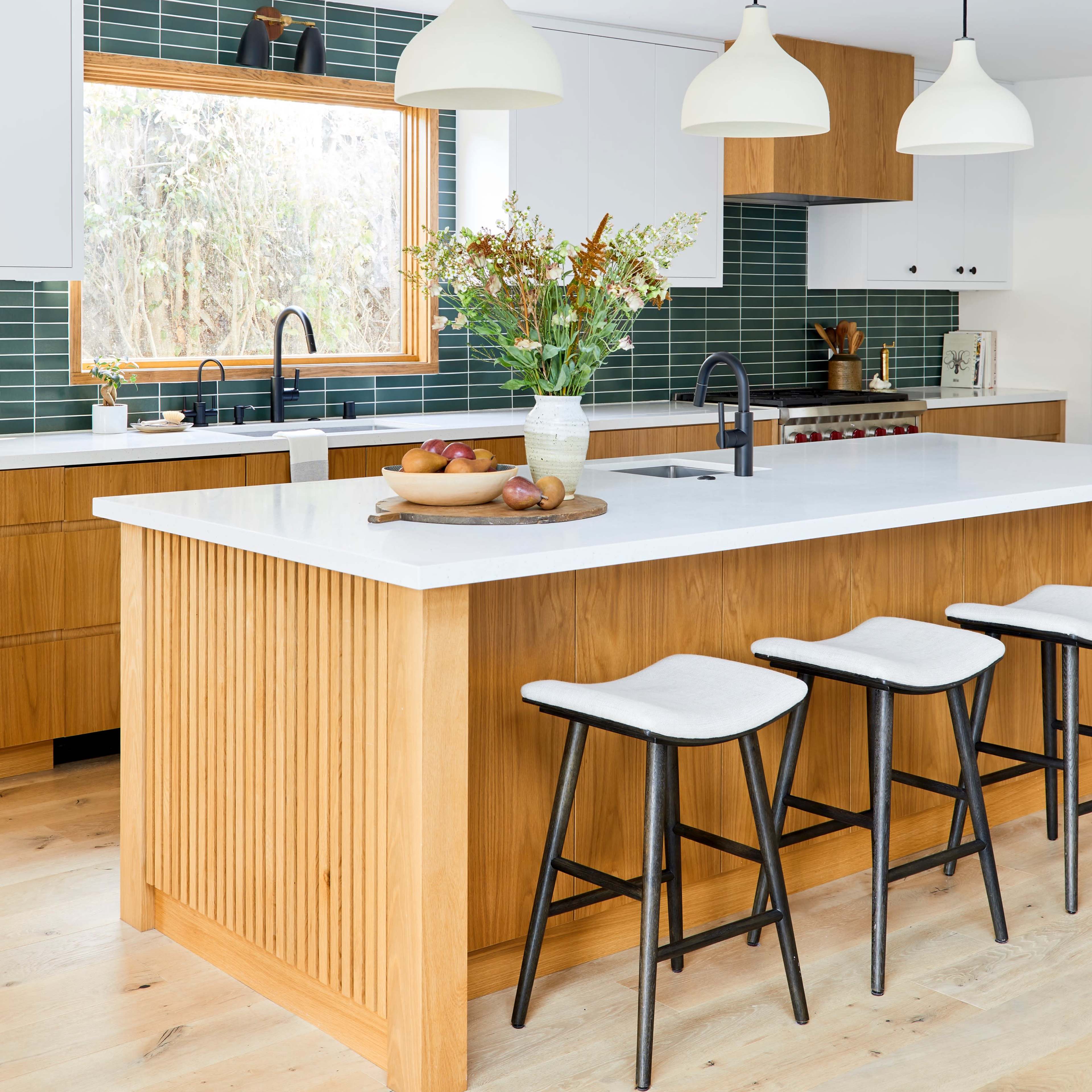 A modern kitchen with a large central island, wooden cabinetry, green tile backsplash, and three pendant lights above.
