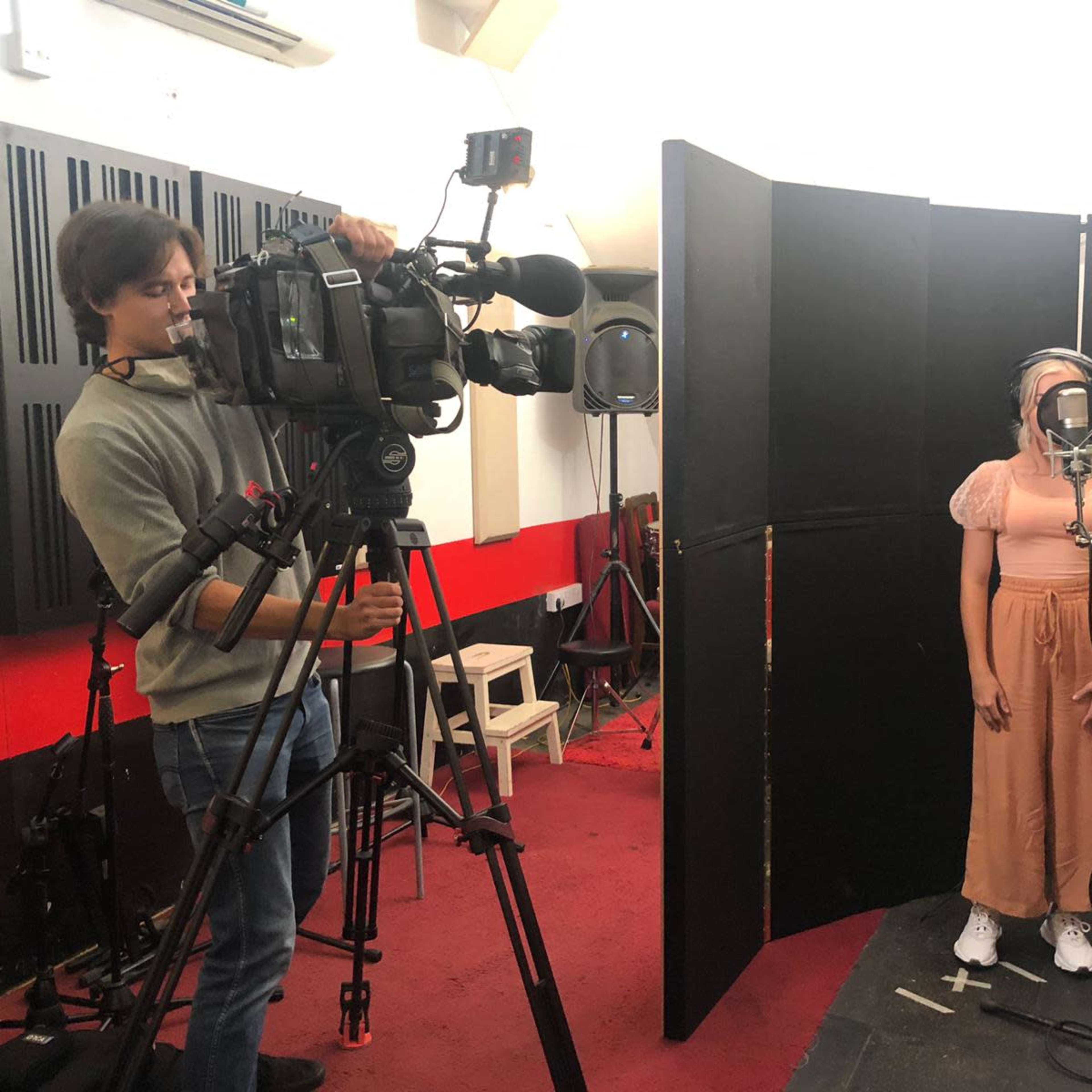 A person stands in front of a microphone in a recording studio while a cameraman films them.