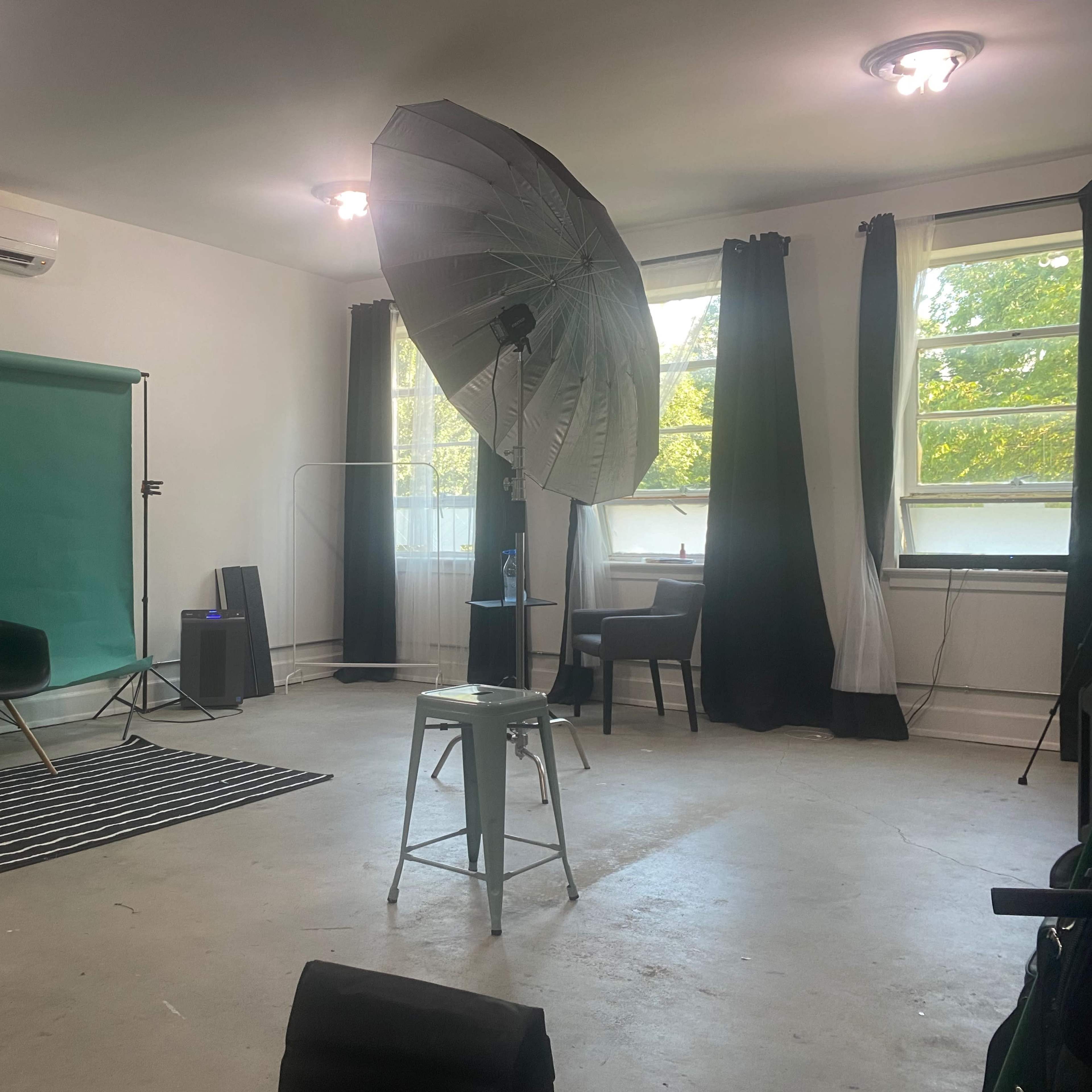 A photography studio featuring a green backdrop, a large softbox light, and minimalist furniture including a chair and a stool.