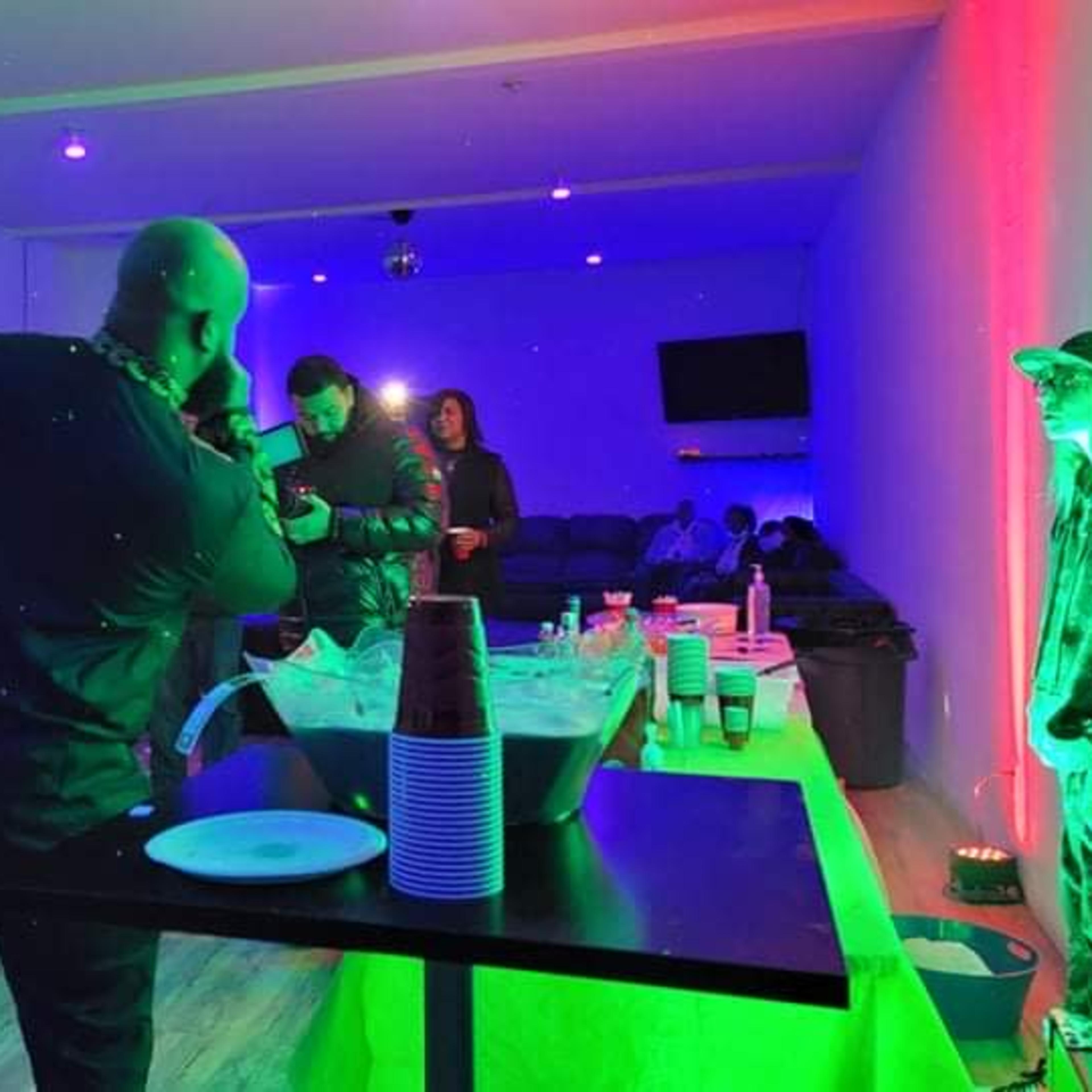 A group of people are gathered in a brightly lit room, with a table displaying drinks and snacks, while others take photos and enjoy the setting.