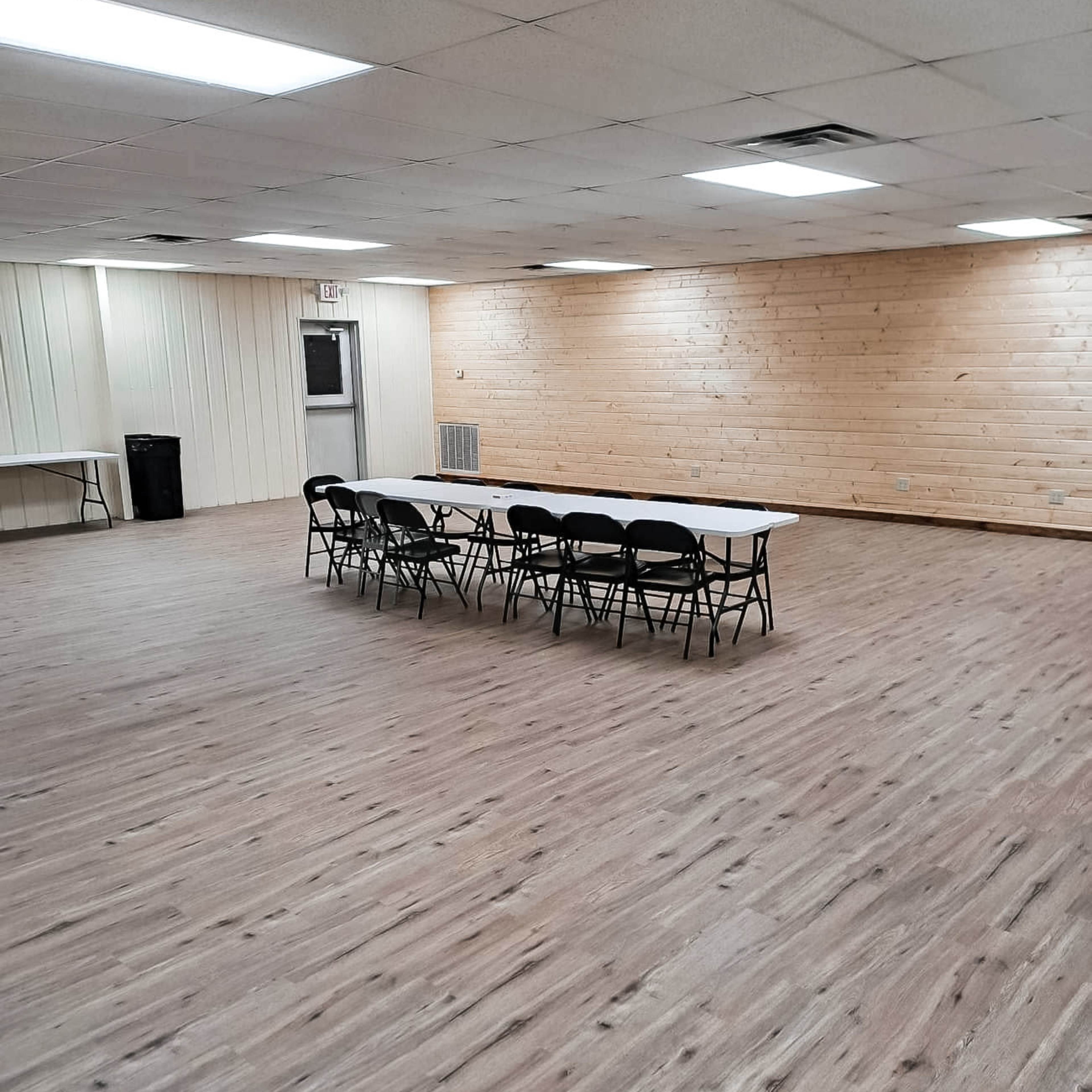 A large room features a long table surrounded by chairs, with wooden walls and a spacious, uncarpeted floor.
