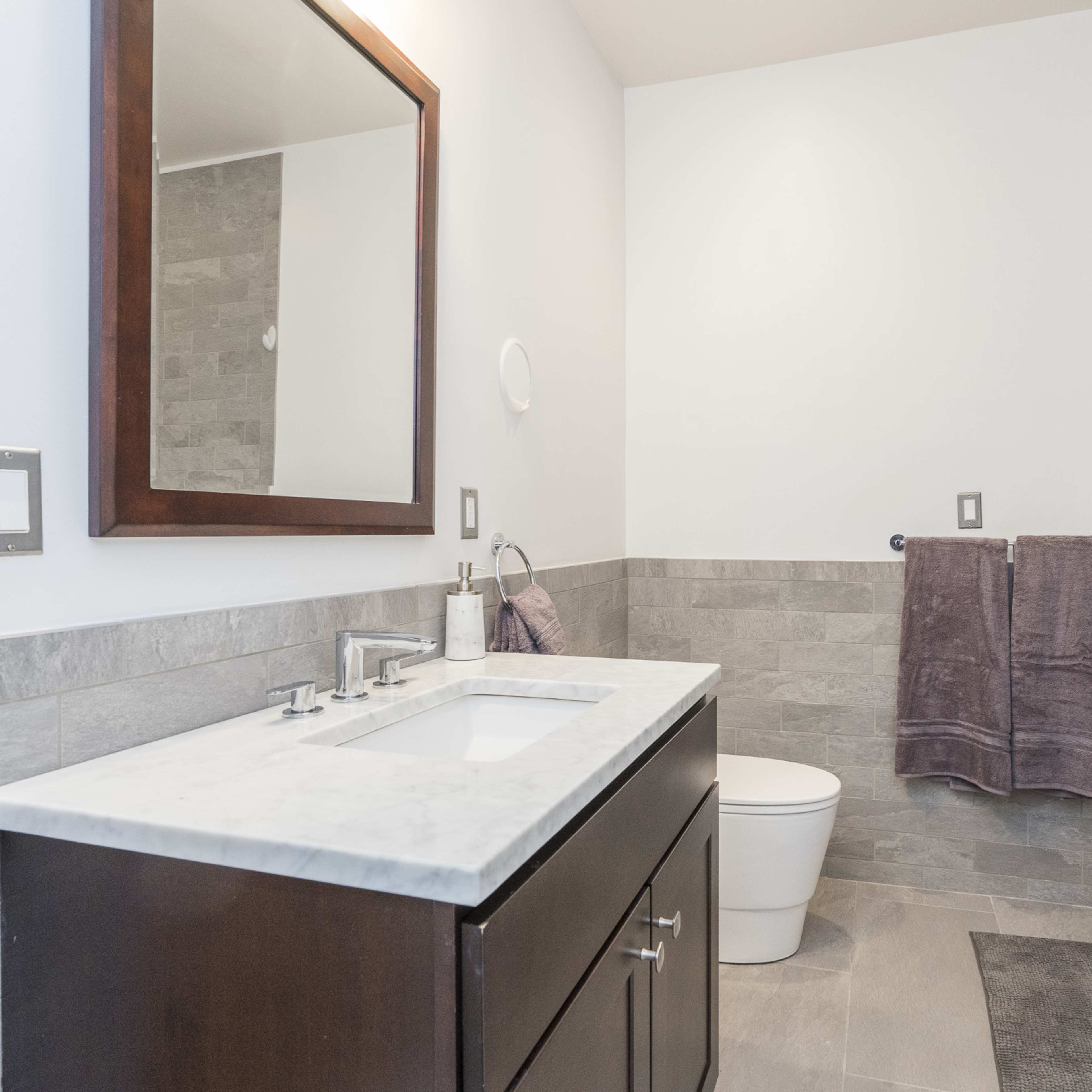 A modern bathroom features a wooden vanity with a marble countertop, a mirror, a toilet, and a glass shower enclosure against a tiled wall.