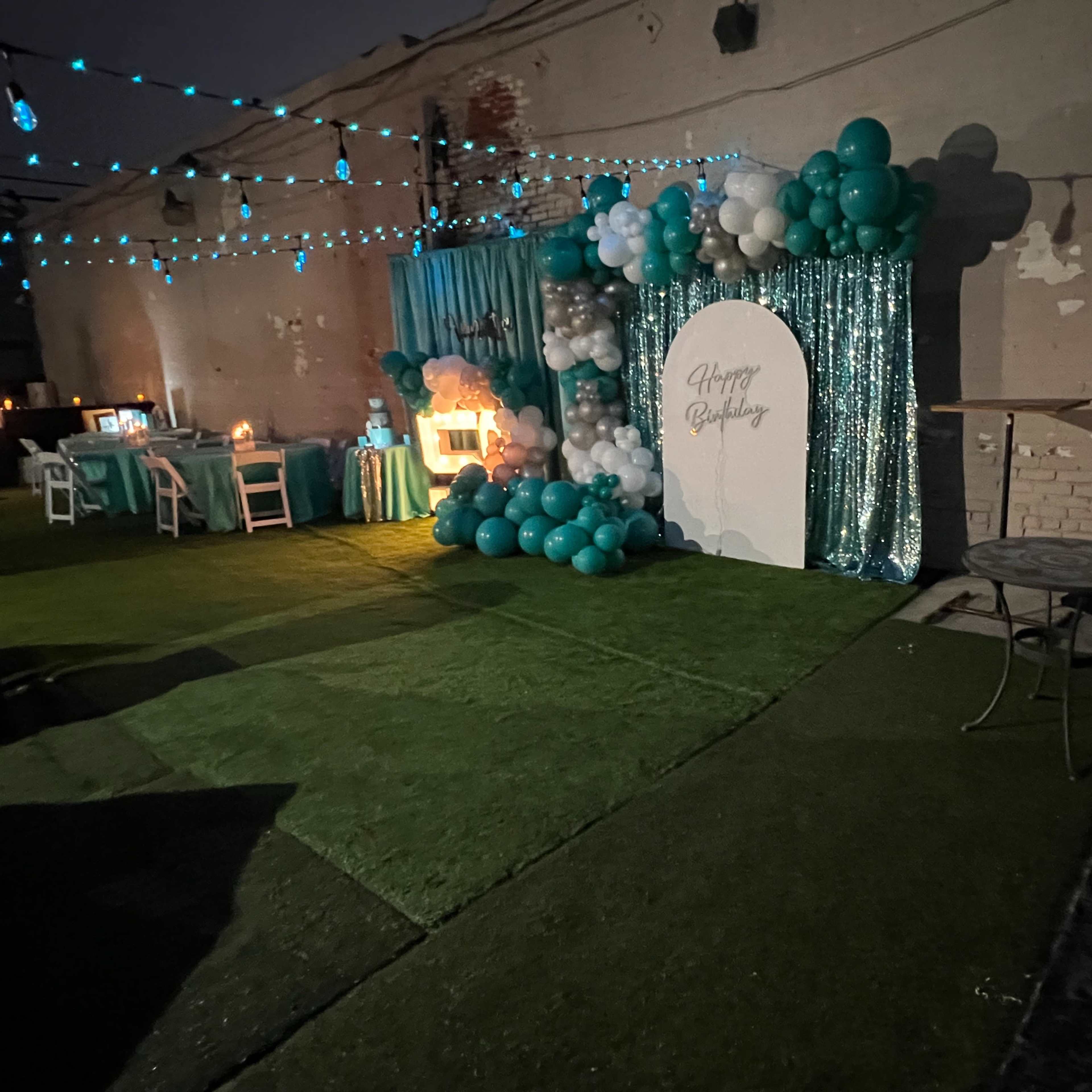 A decorated outdoor event space with a backdrop featuring balloons and a "Happy Birthday" sign, illuminated by string lights.